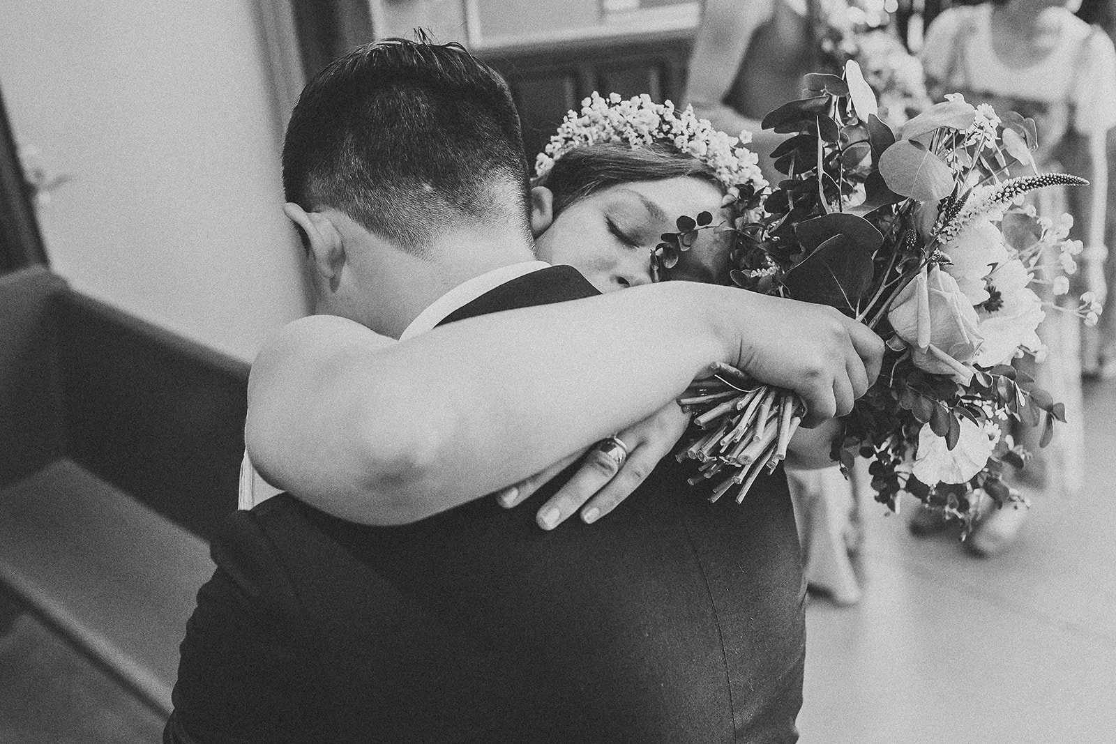 A couple hugging, with the woman holding a bouquet of flowers, in a black and white photo.