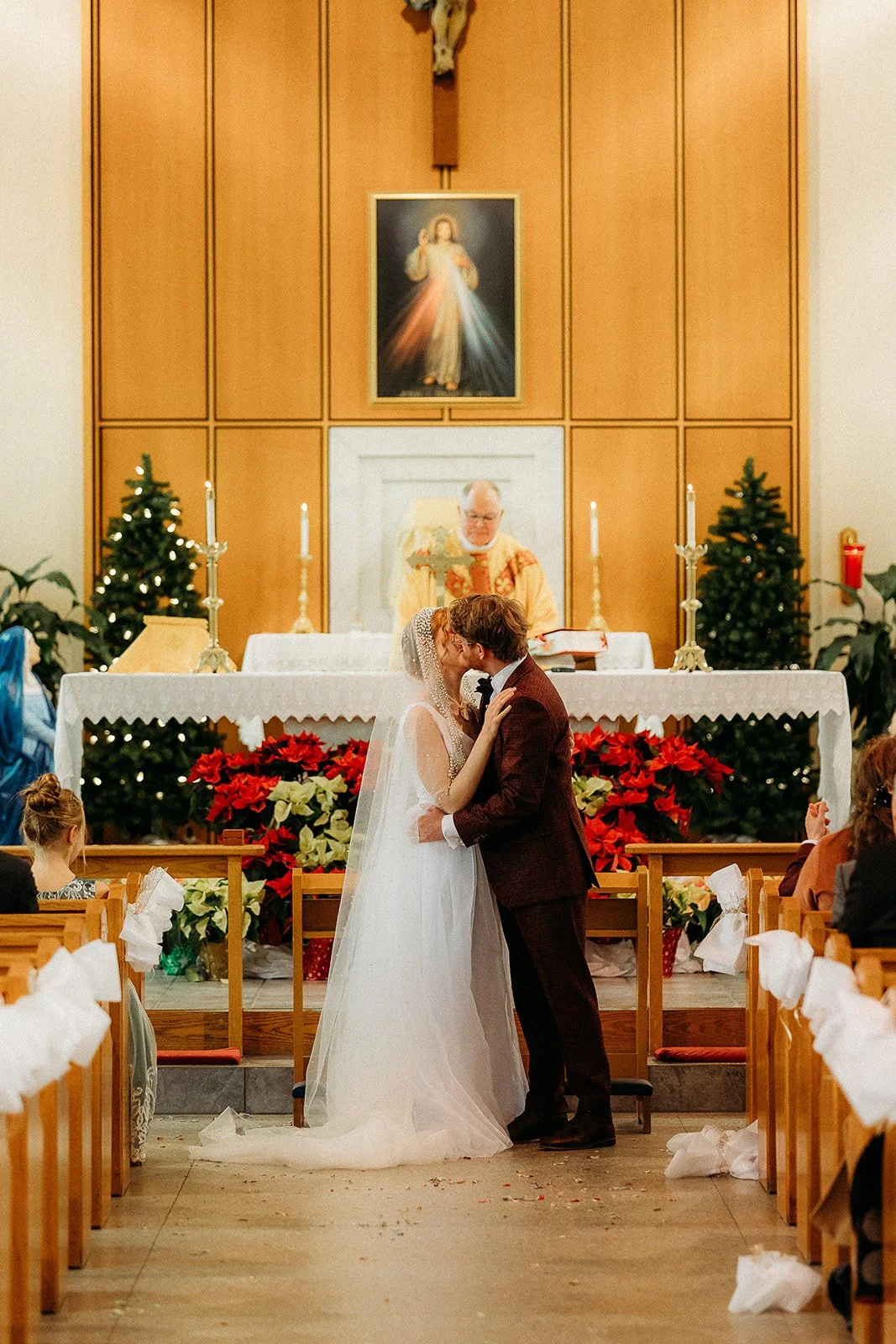 A wedding ceremony inside a church with a bride and groom kissing at the altar, priest standing behind them, Christmas decorations, and a crucifix and religious painting on the wooden wall behind.