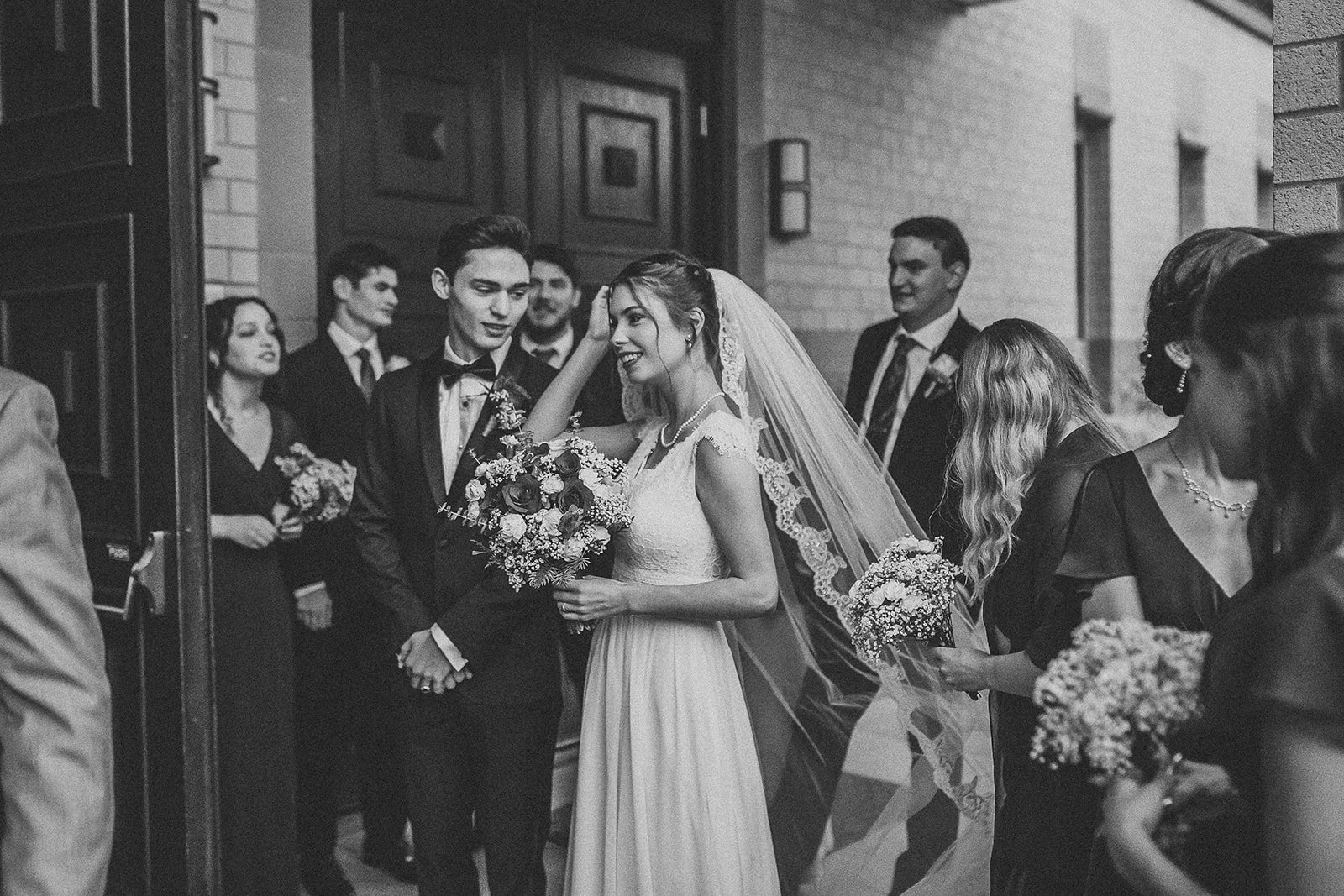 Black and white photograph of a wedding reception with a bride and groom surrounded by friends and family outside a building, the bride holding a bouquet of flowers and wearing a lace wedding dress with a veil, while the groom is in a tuxedo, and oth