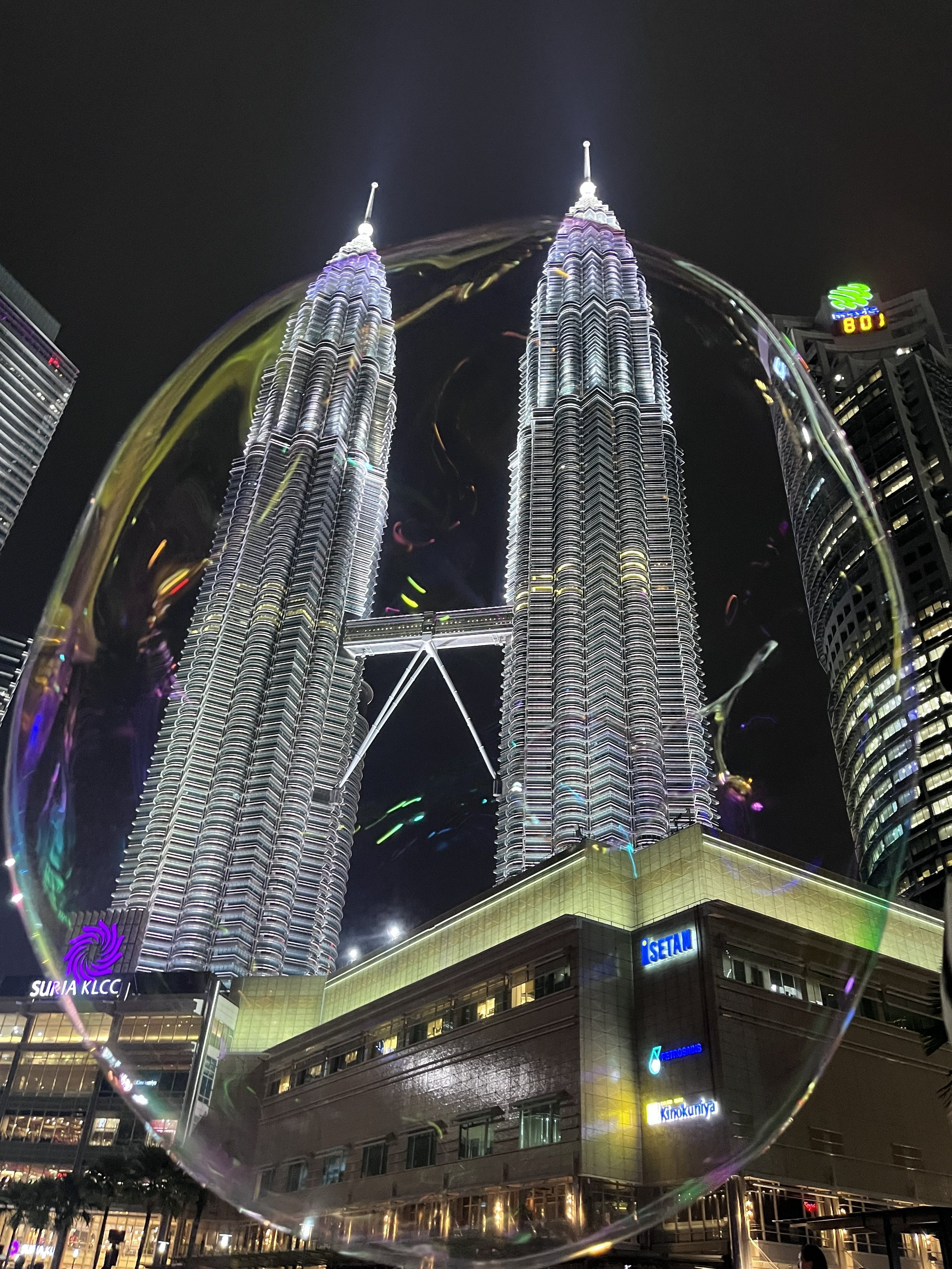 Nighttime view of the Petronas Towers in Kuala Lumpur, Malaysia, seen through a wine glass. The towers are illuminated and reflect on the glass, with surrounding buildings also lit up.