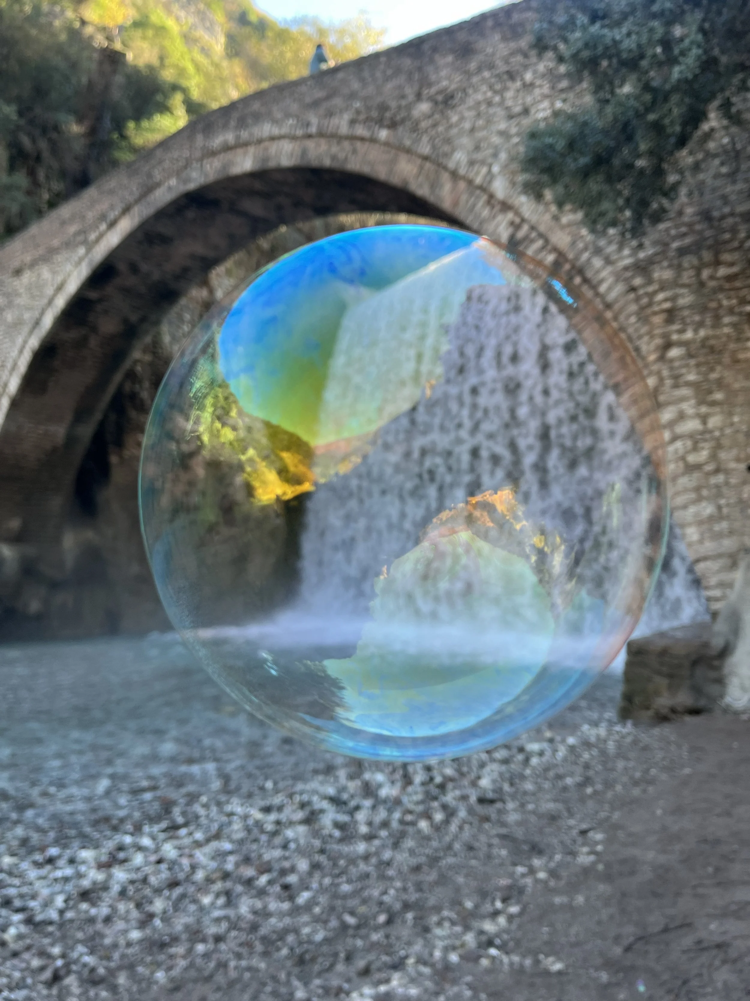 A clear soap bubble reflecting the sky, trees, and surroundings, floating in front of a stone arched bridge over a gravel pathway.
