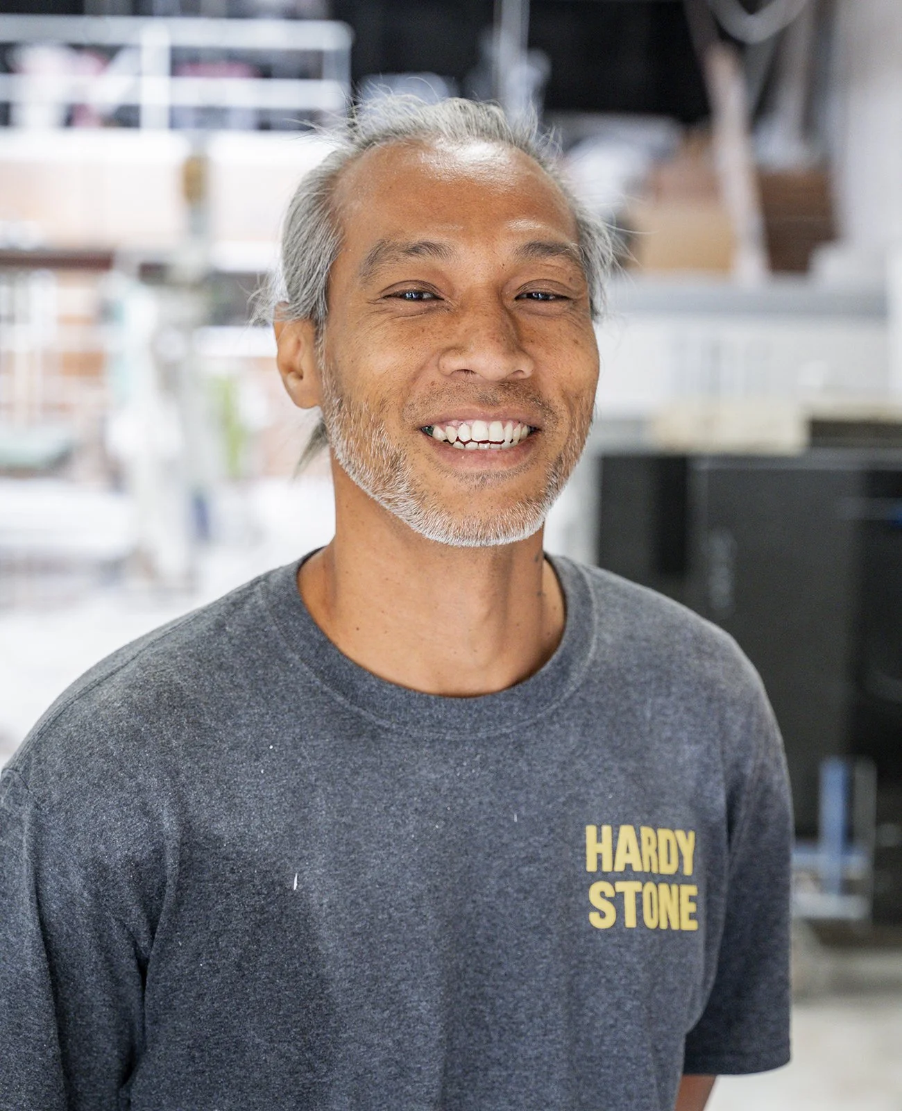 Smiling man in gray "Hardy Stone" T-shirt inside a workshop.