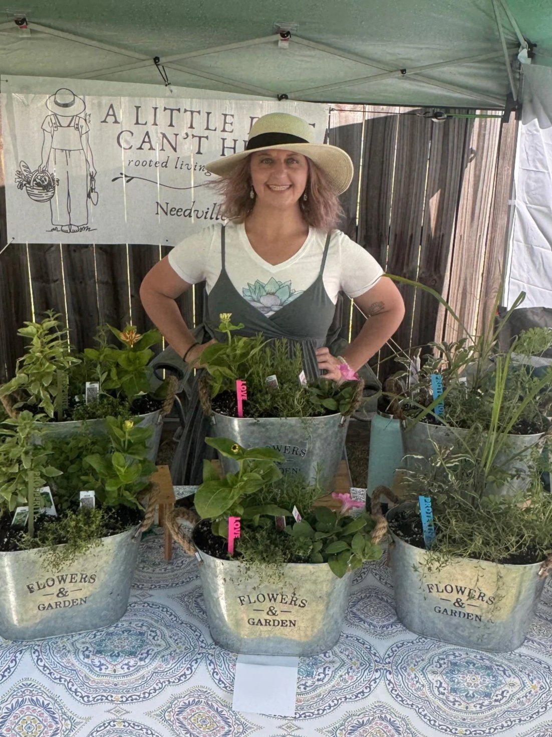 Nicole from A Little Dirt Can't Hurt is wearing a wide-brimmed garden hat and overalls with a floral design. She's standing behind a table with potted plants at a garden market booth.