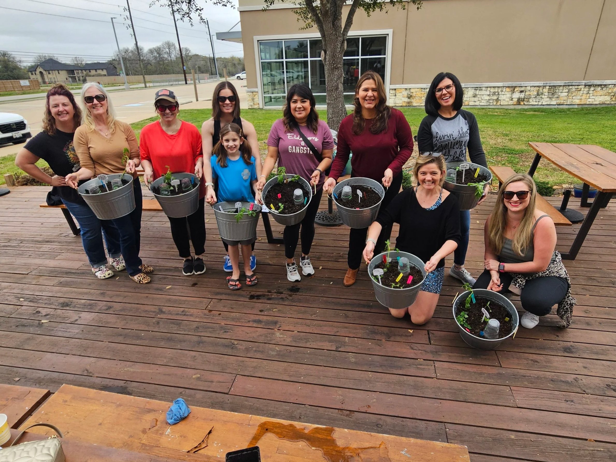 A group of women and a young girl smiling and holding buckets with small plants inside, standing on a wooden deck outdoors after attending a workshop with Nicole at A Little Dirt Can't Hurt
