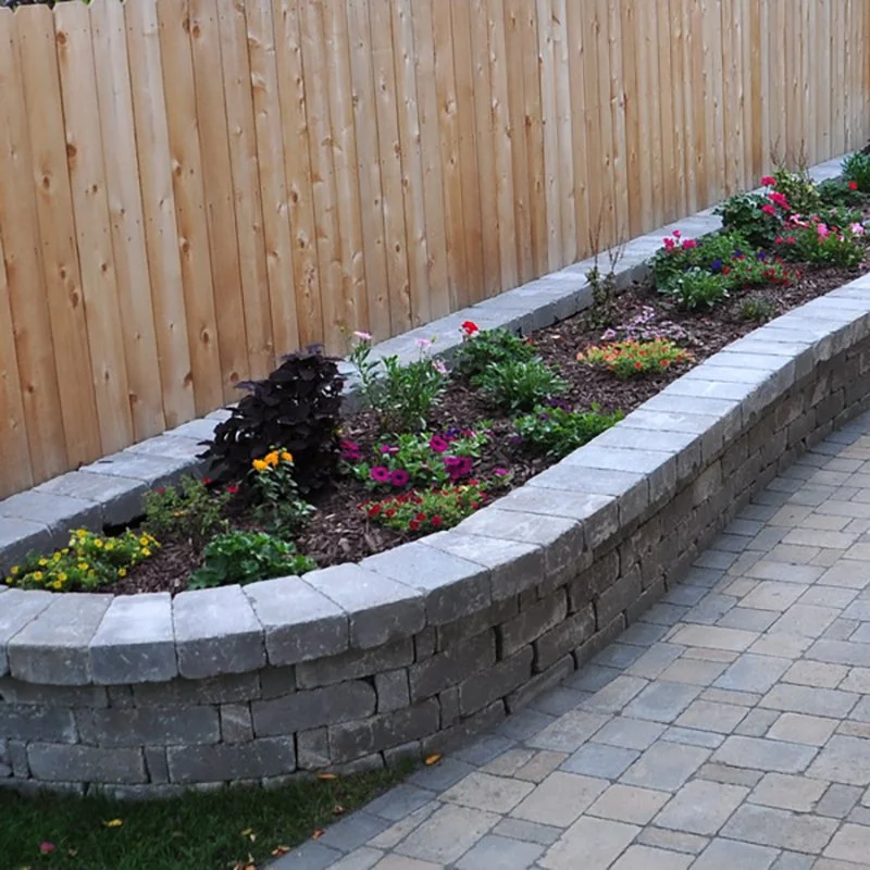 A curved garden bed with colorful flowers, bordered by gray bricks, next to a wooden fence and paved walkway.