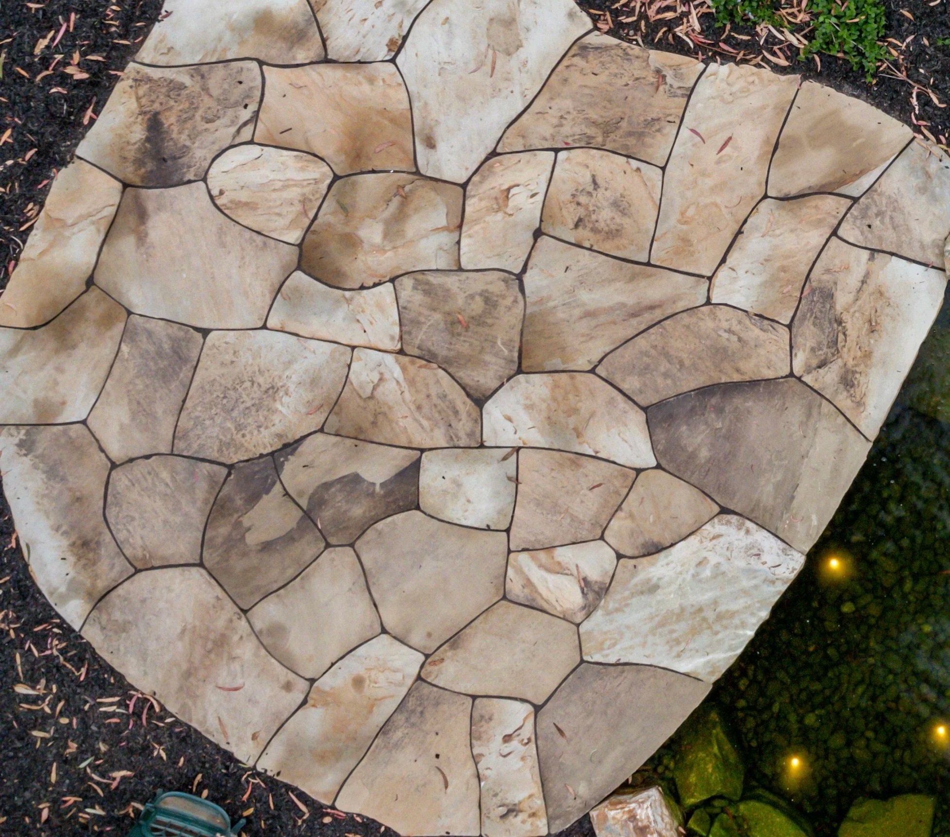 Stone pathway with irregularly shaped beige and brown stones, surrounded by black gravel and a water feature with lily pads and floating lights.
