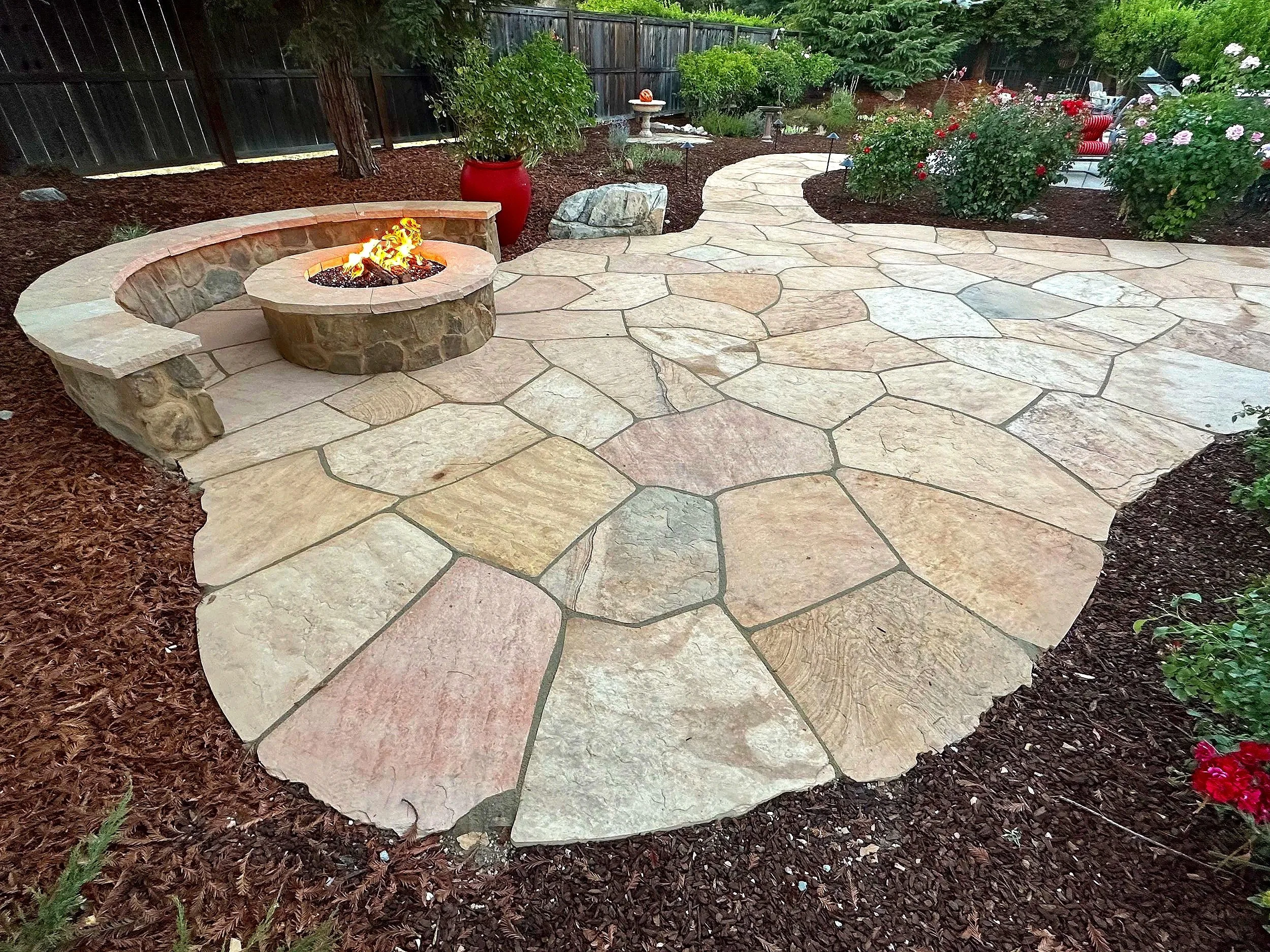 A backyard patio with flagstone flooring, a fire pit with a stone surround, and garden beds with blooming roses and potted plants.