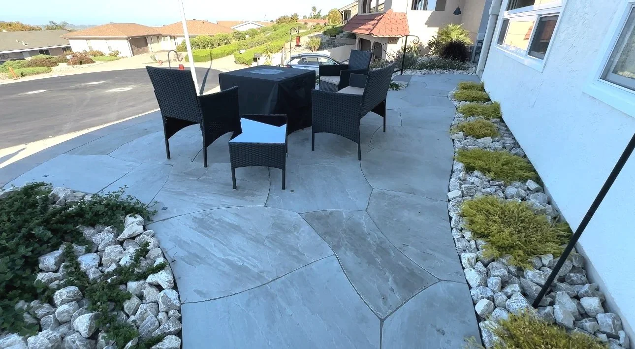 A curved patio with stone paving, outdoor black wicker chairs, and a table covered with a black cloth. The patio is bordered by white rocks and yellow-green plants, with a house wall on the right side and a suburban street in the background.