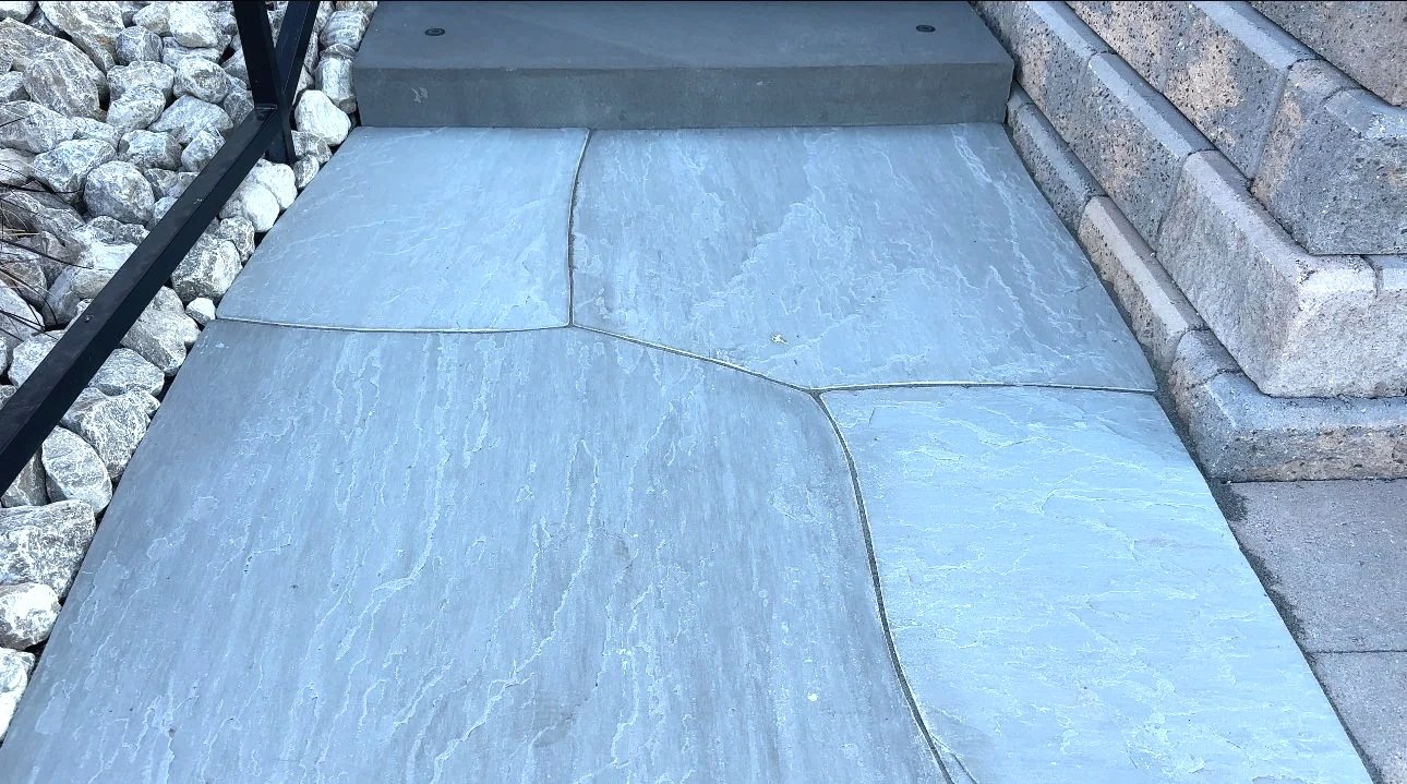 Close-up of decorative gray concrete sidewalk with visible texture, bordered by stacked stone on the right and gravel with a fence on the left.