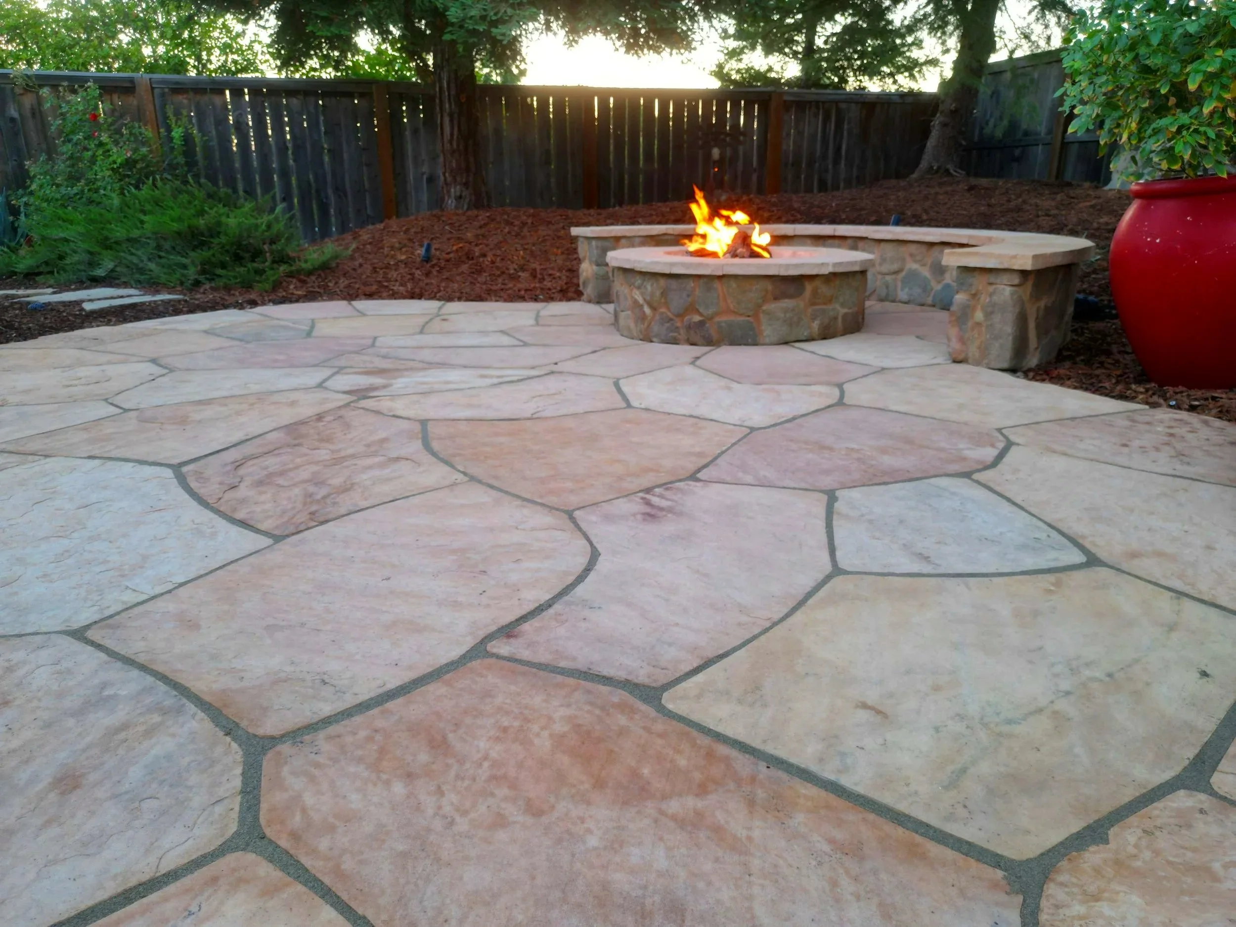 A backyard patio with large irregular stone pavers, a stone fire pit with a small fire, and a red large ceramic pot on the right side, with a wooden fence and trees in the background.