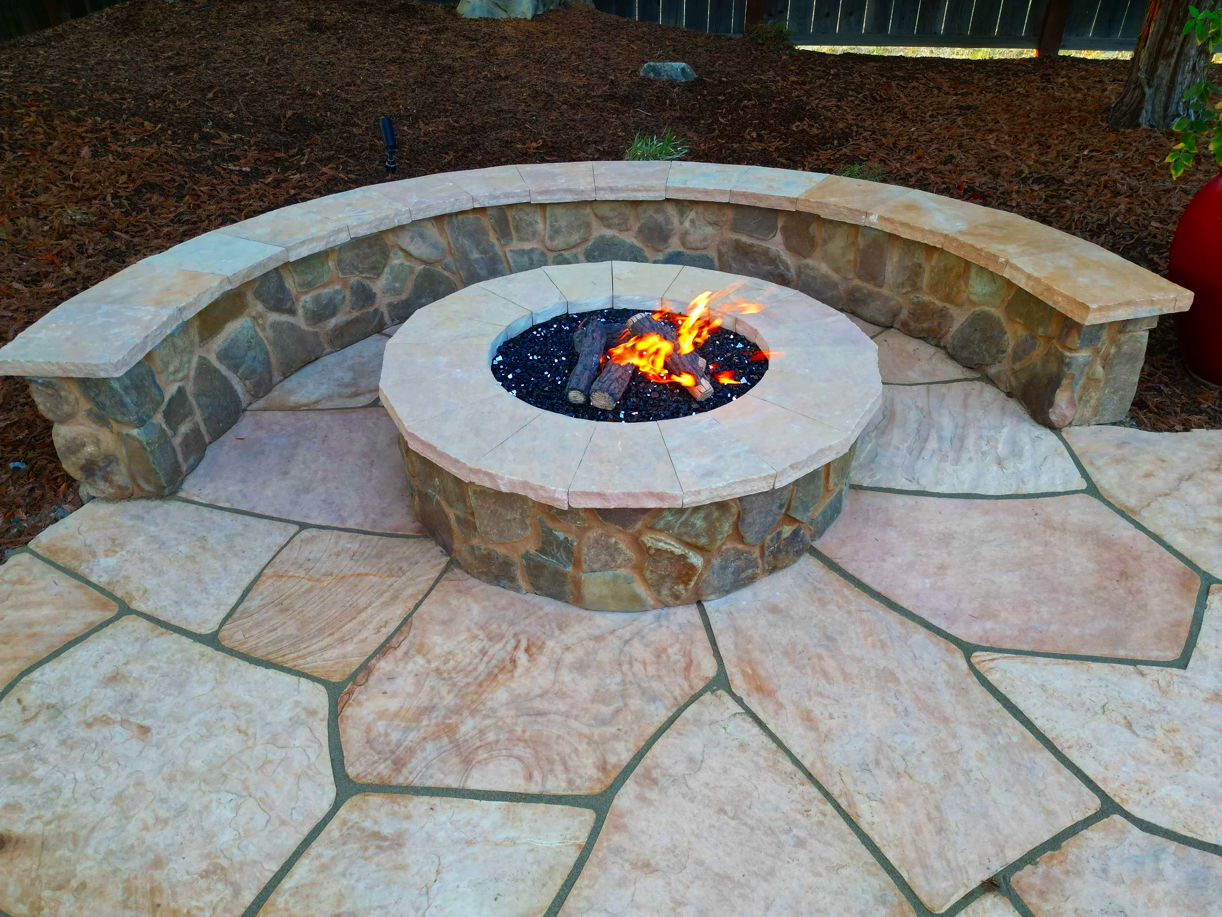 Stone fire pit with a circular seating wall and a seating bench, set on large flagstone pavers, outdoors with mulch and trees in the background.
