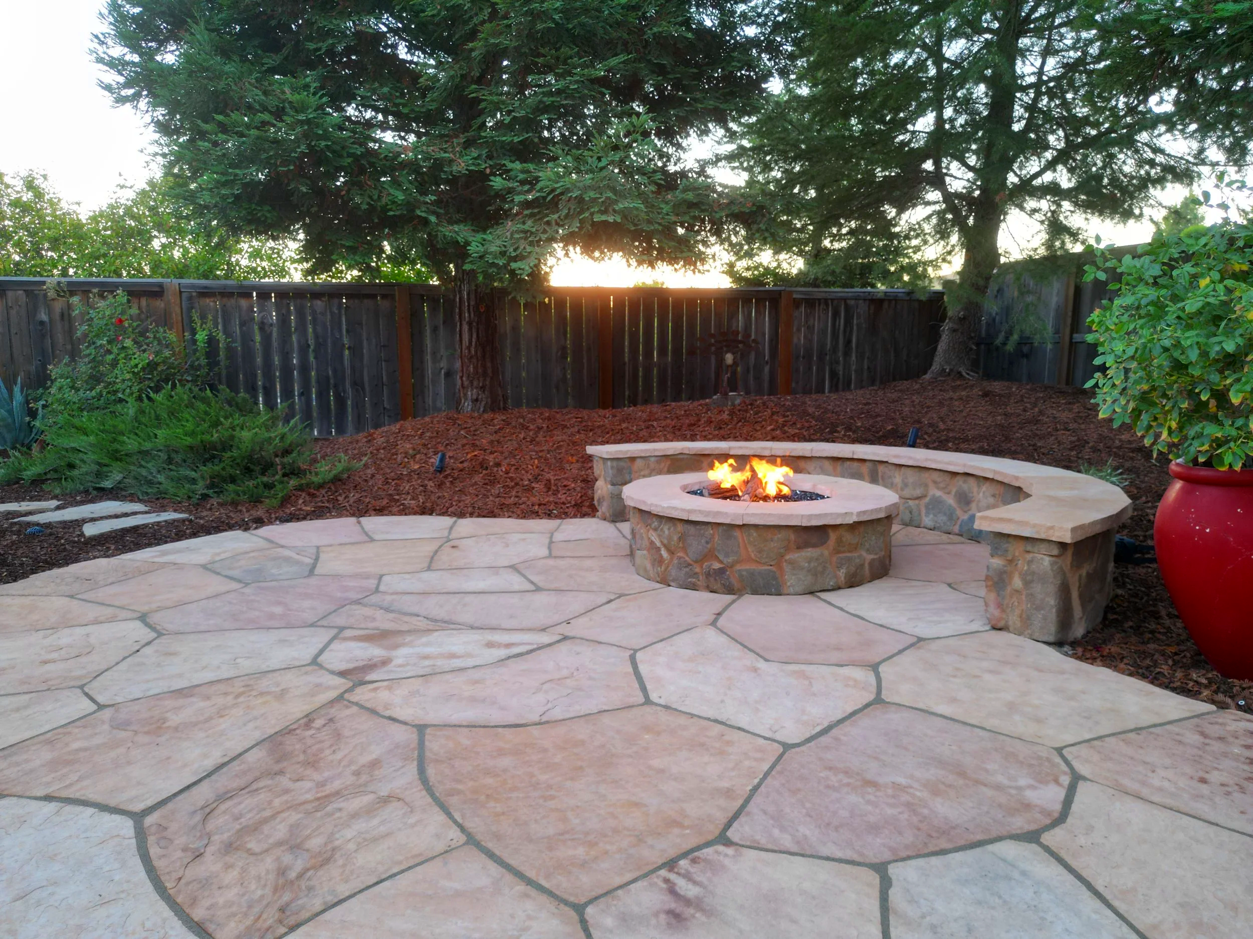 Backyard patio with a stone fire pit and seating area, surrounded by a wooden fence, trees, and various plants.