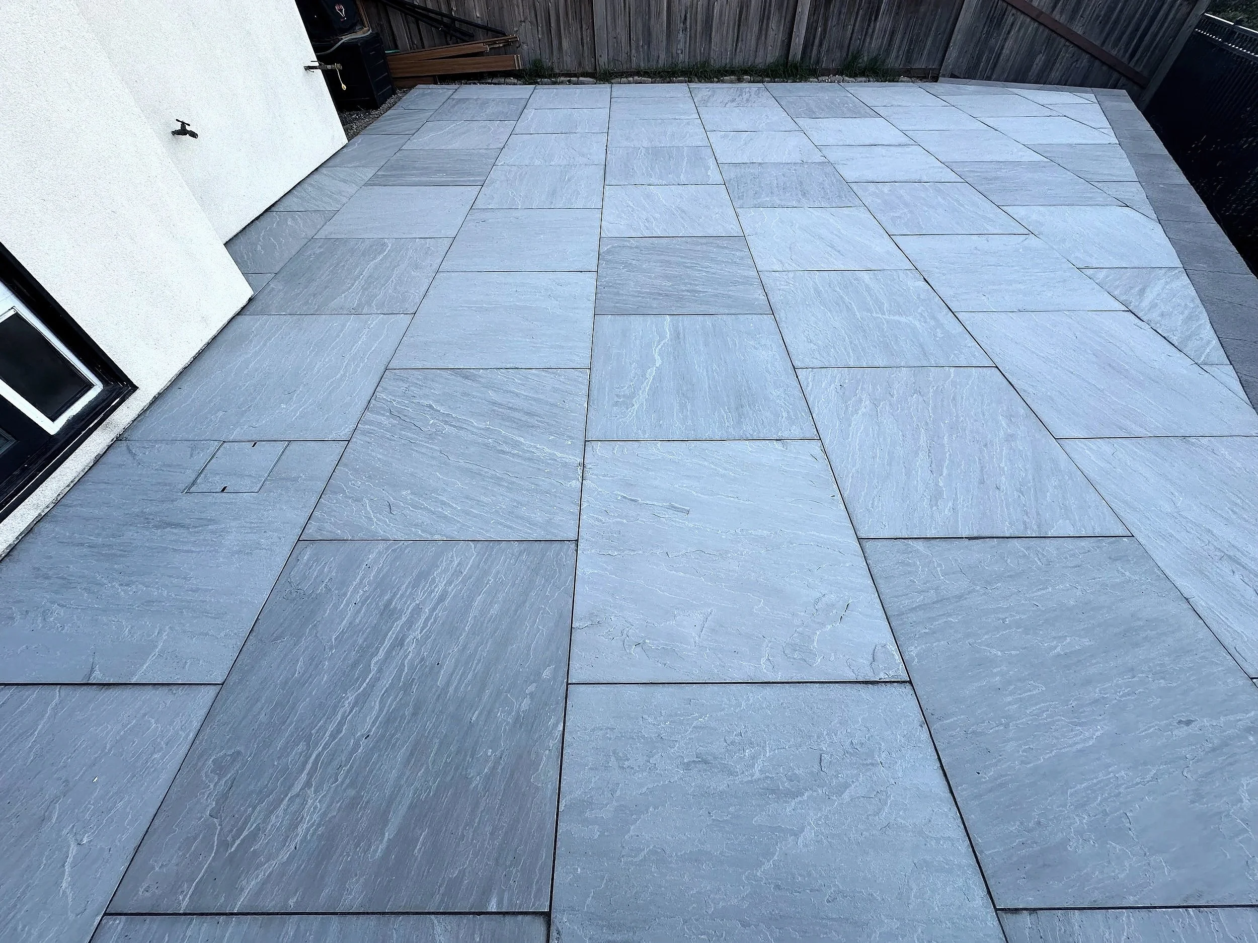 An outdoor patio with large gray slate tiles, adjacent to a white exterior wall and a black privacy fence.