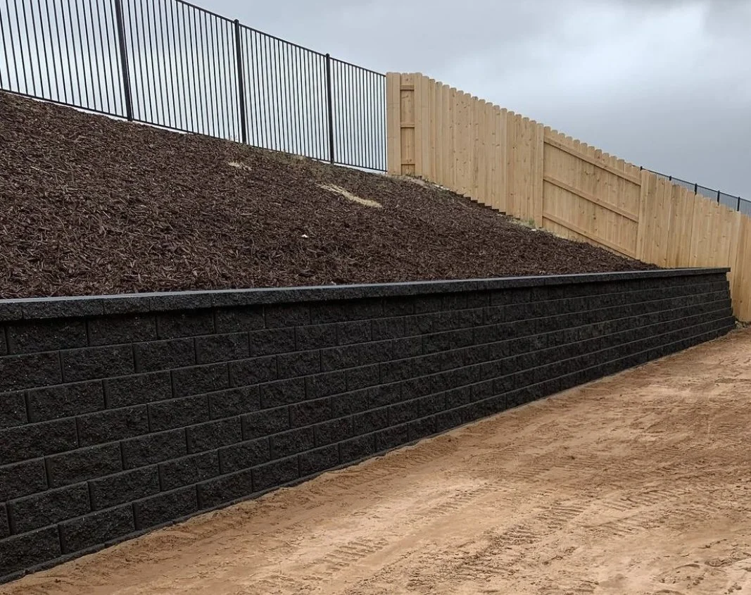 A landscaped hillside shows a black retaining wall at the bottom, a layer of mulch above it, a wooden fence on the hillside, and a black metal fence at the top of the hill, under an overcast sky.
