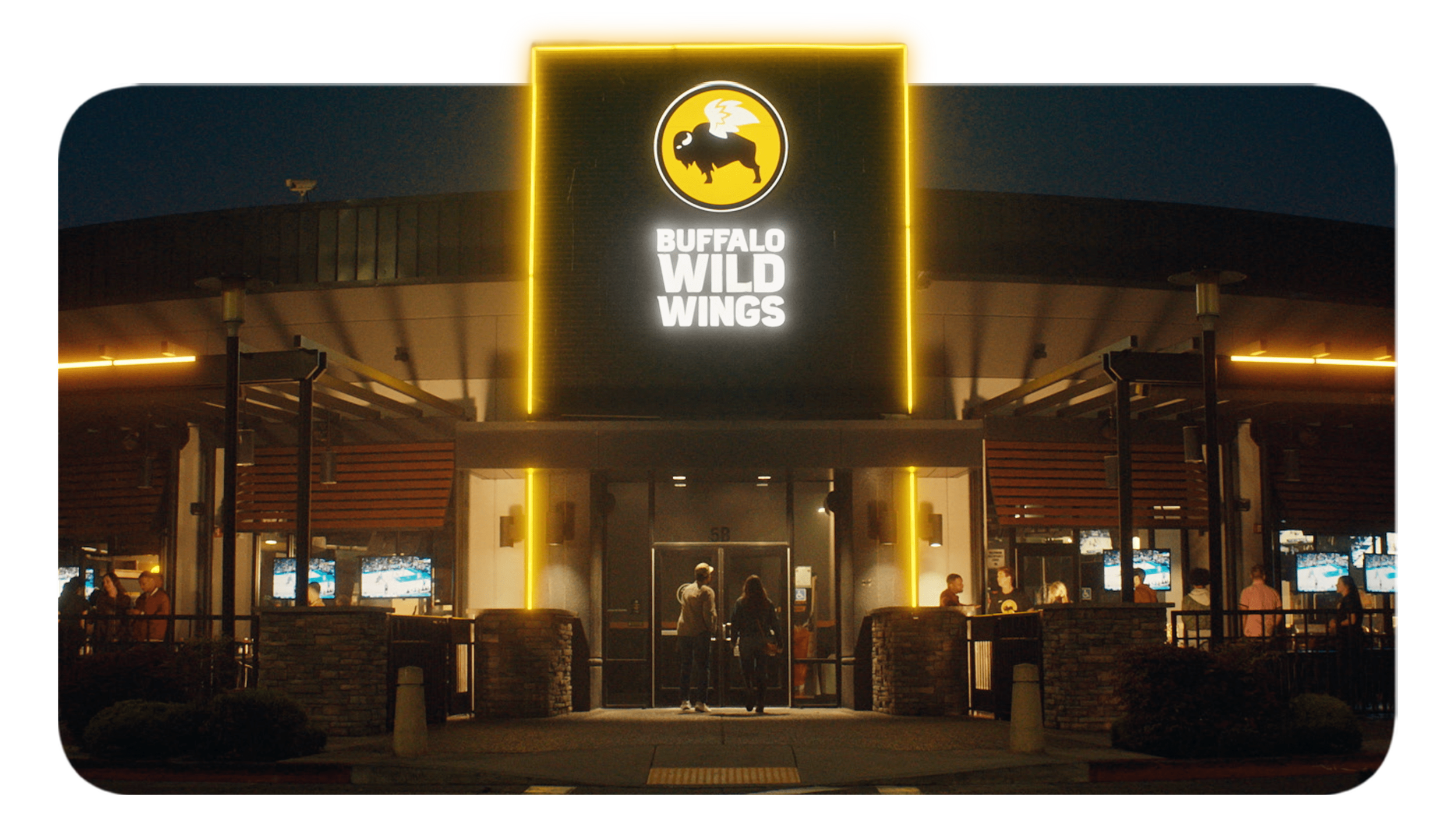 Night view of Buffalo Wild Wings restaurant entrance with illuminated sign featuring a buffalo with lightning wings. People are entering and standing outside, with multiple screens visible inside showing sports.