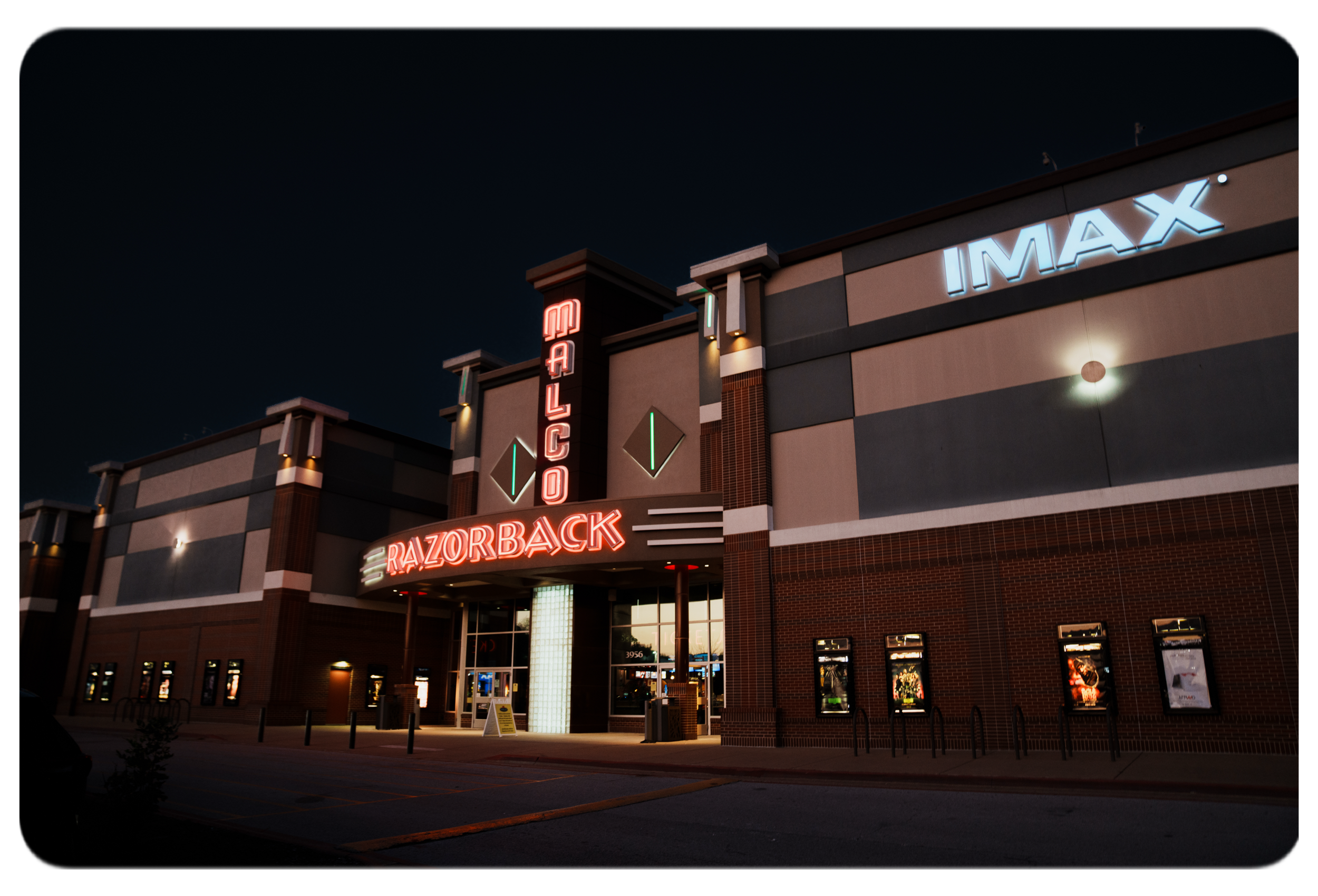 Night view of a movie theater with illuminated signs for 'Malco Razorsback' and 'IMAX'. The building has a modern exterior with brick and concrete walls, and the entrance is lit up with neon lights.