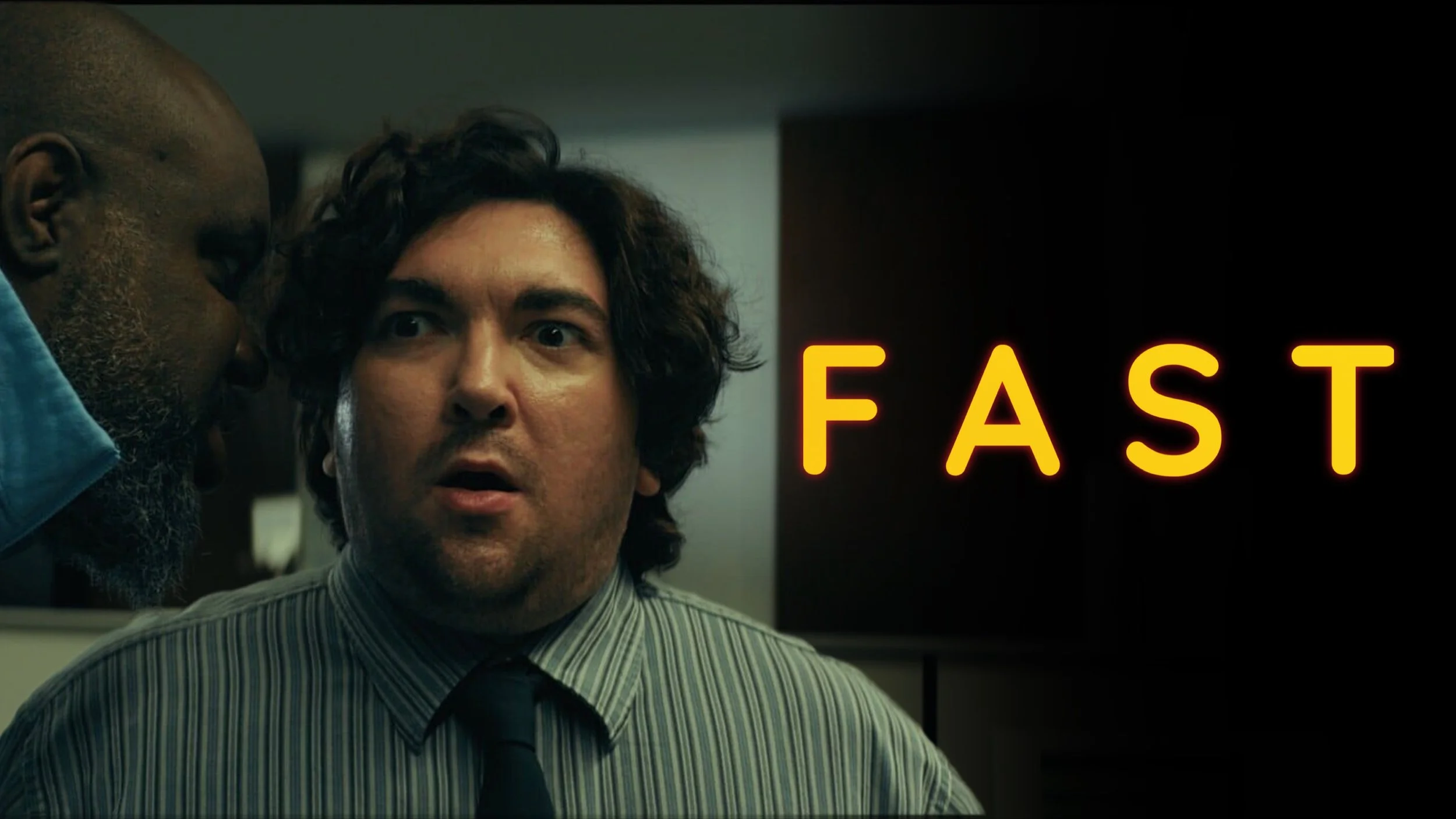 A man with dark hair and a striped shirt is looking surprised or shocked while another man, with gray hair and beard, appears to whisper into his ear. The background features a lit-up black sign with yellow-orange text that reads 'FAST'.