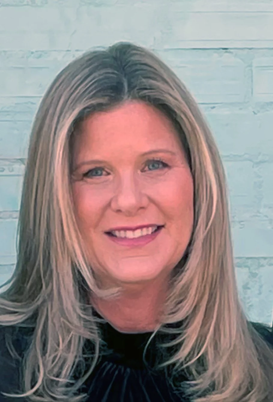 Portrait of a woman with shoulder-length blonde hair, smiling, in front of a light-colored brick wall background.