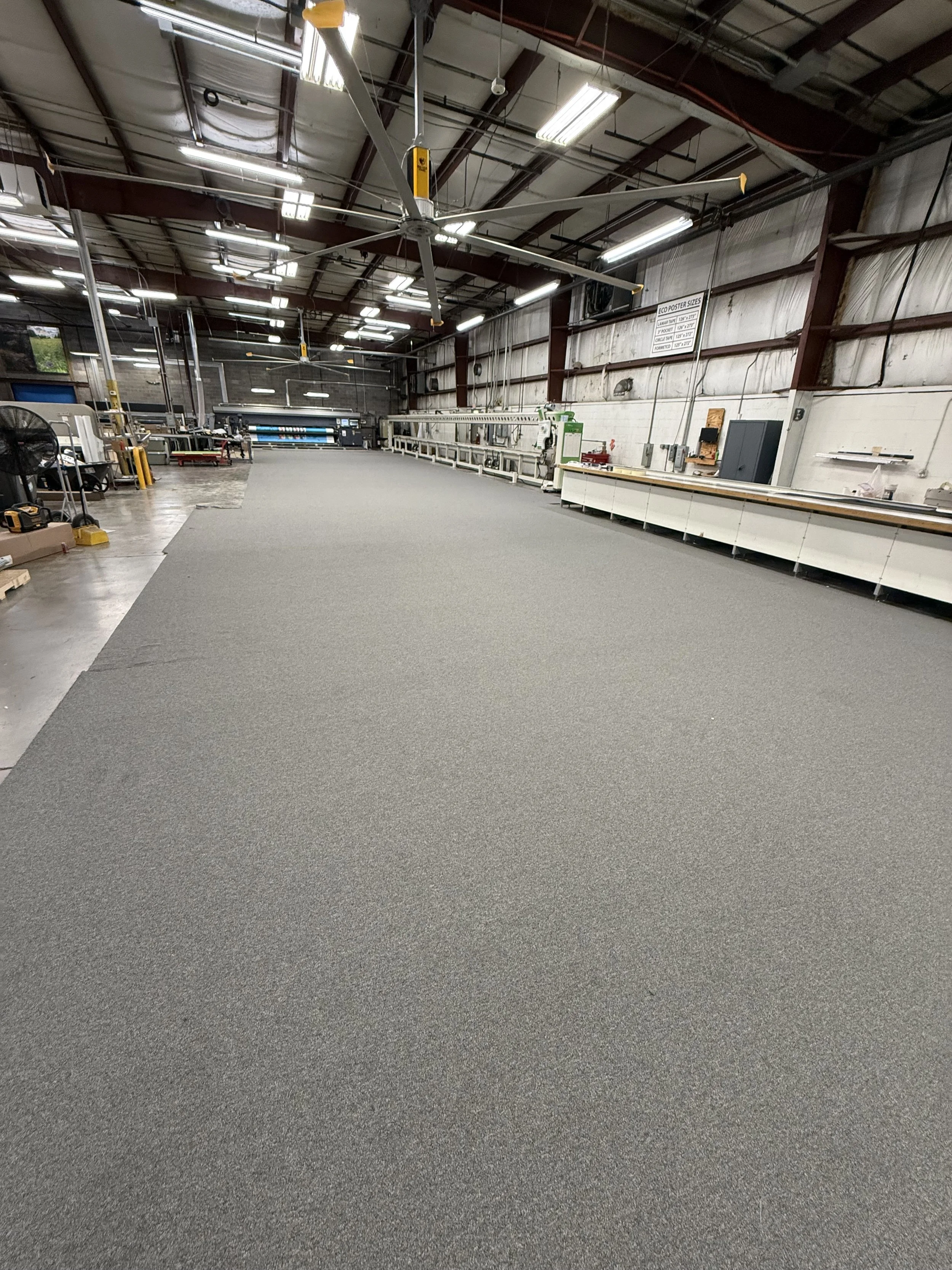 Empty industrial warehouse with overhead fans, fluorescent lights, workstations, and a large gray carpeted area in the center.