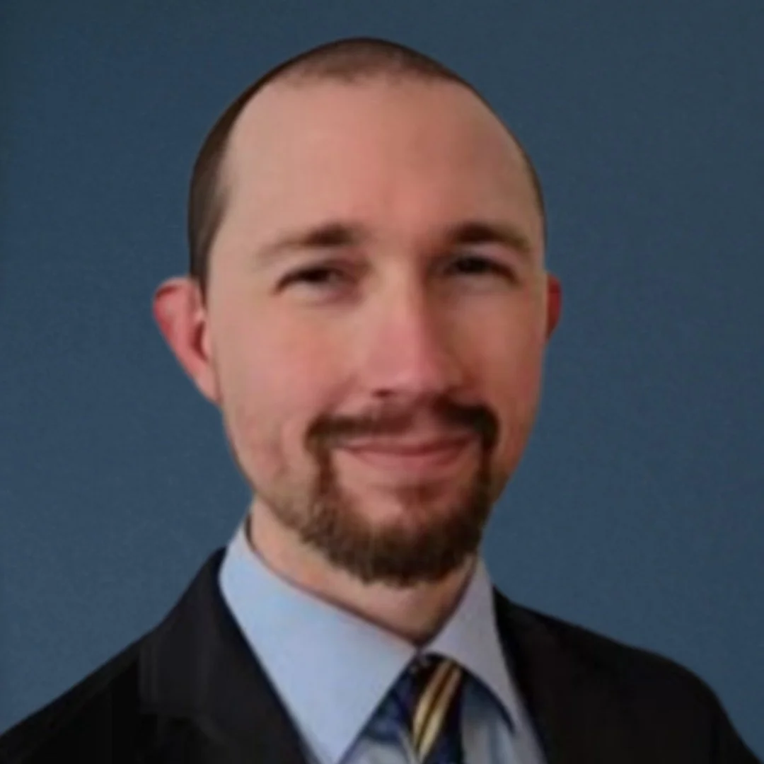 A man with short brown hair, a beard, and mustache, dressed in a dark suit and a striped tie, smiling against a blue background.