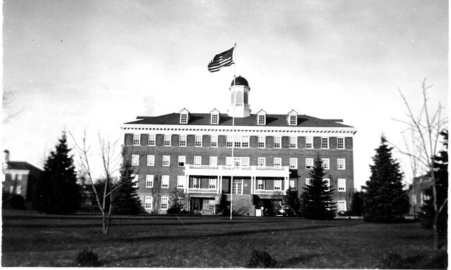 Black and white historic photo of a large brick hotel with an American flag and another flag flying from the roof, surrounded by trees and a lawn.