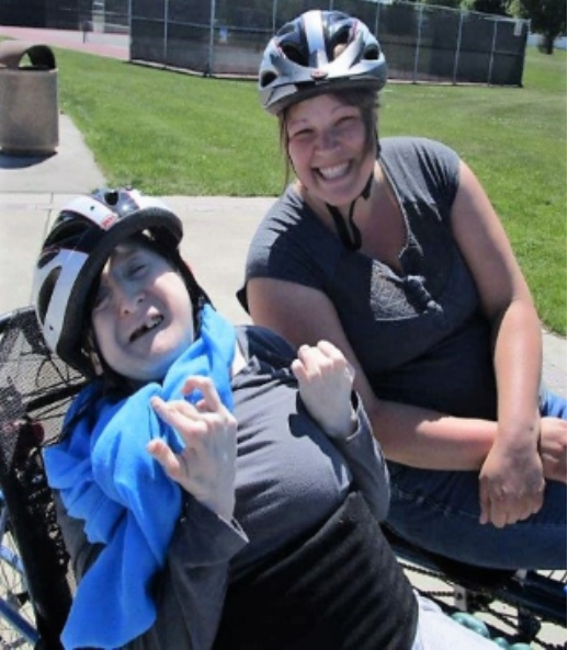 Two smiling individuals wearing helmets, outdoors on a sunny day. One is in a wheelchair with a blue jacket, and the other is sitting beside them on the grass.