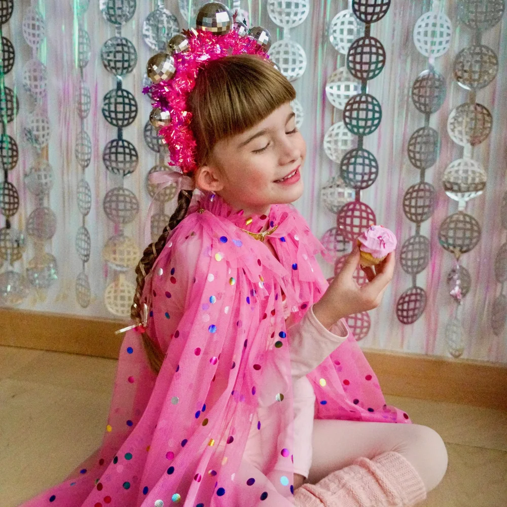 A smiling girl in a pink polka-dot cape and a disco-ball crown holding a frosted cupcake in front of a shimmering disco-ball backdrop
