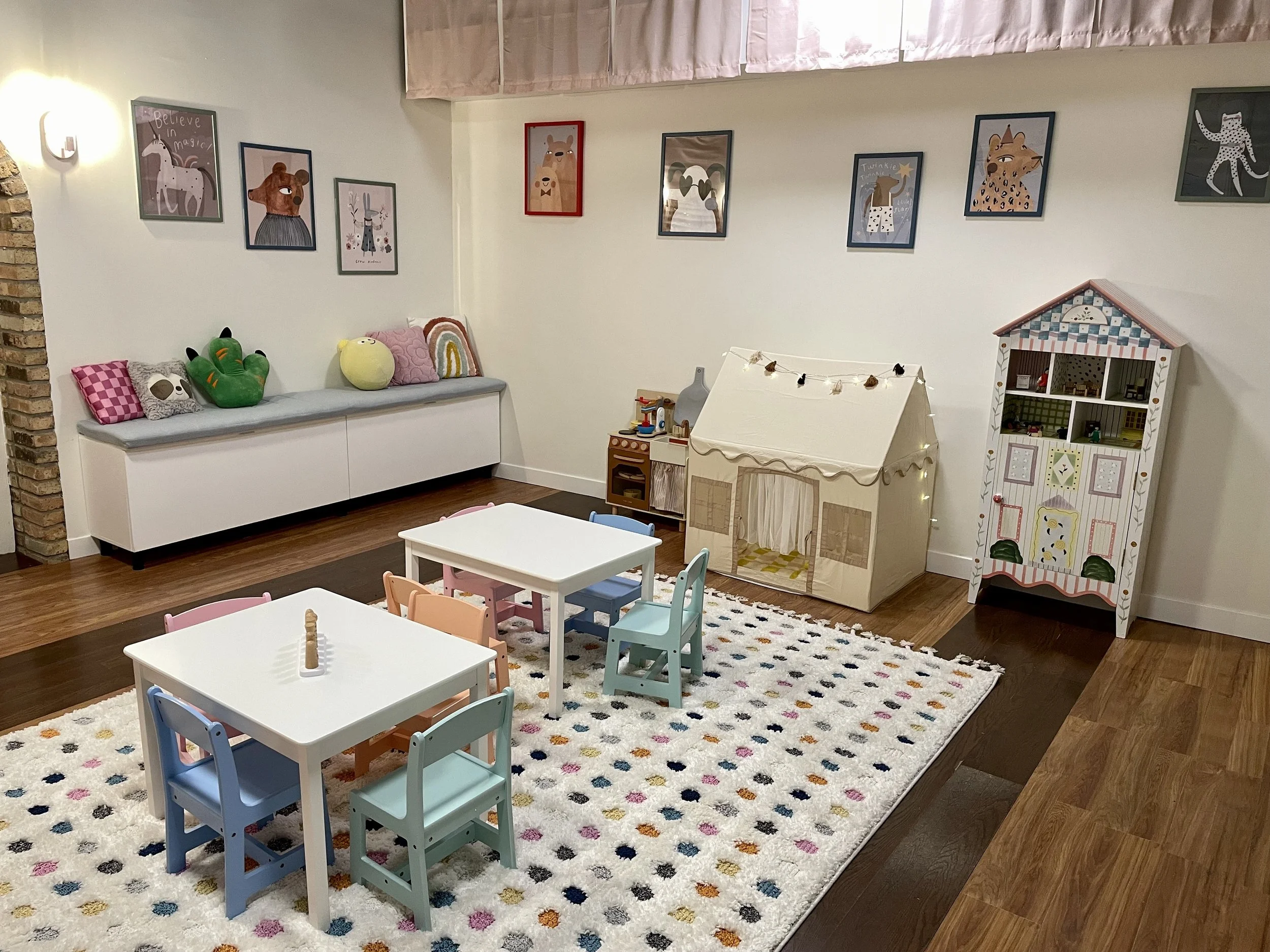 A children's playroom with small tables and chairs, a plush rug with colorful dots, a shelf with stuffed animals, framed animal art on the walls, and a play tent.