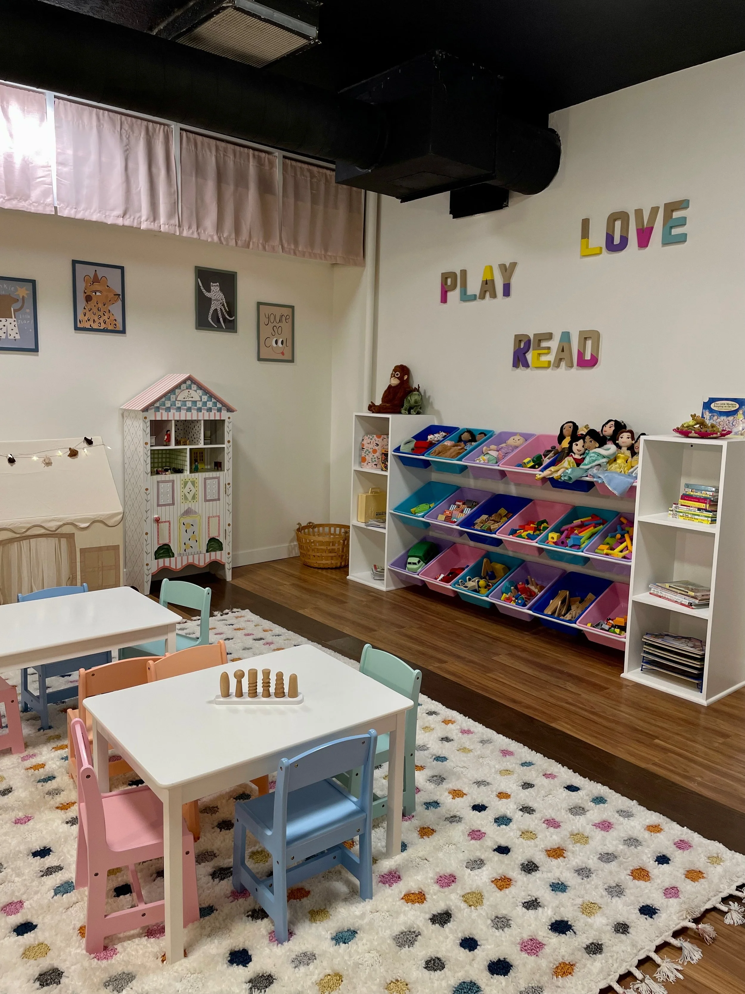 Colorful children's playroom with small tables and chairs, toy shelves filled with toys, framed animal artwork on the wall, and the words 'PLAY LOVE READ' on the wall.