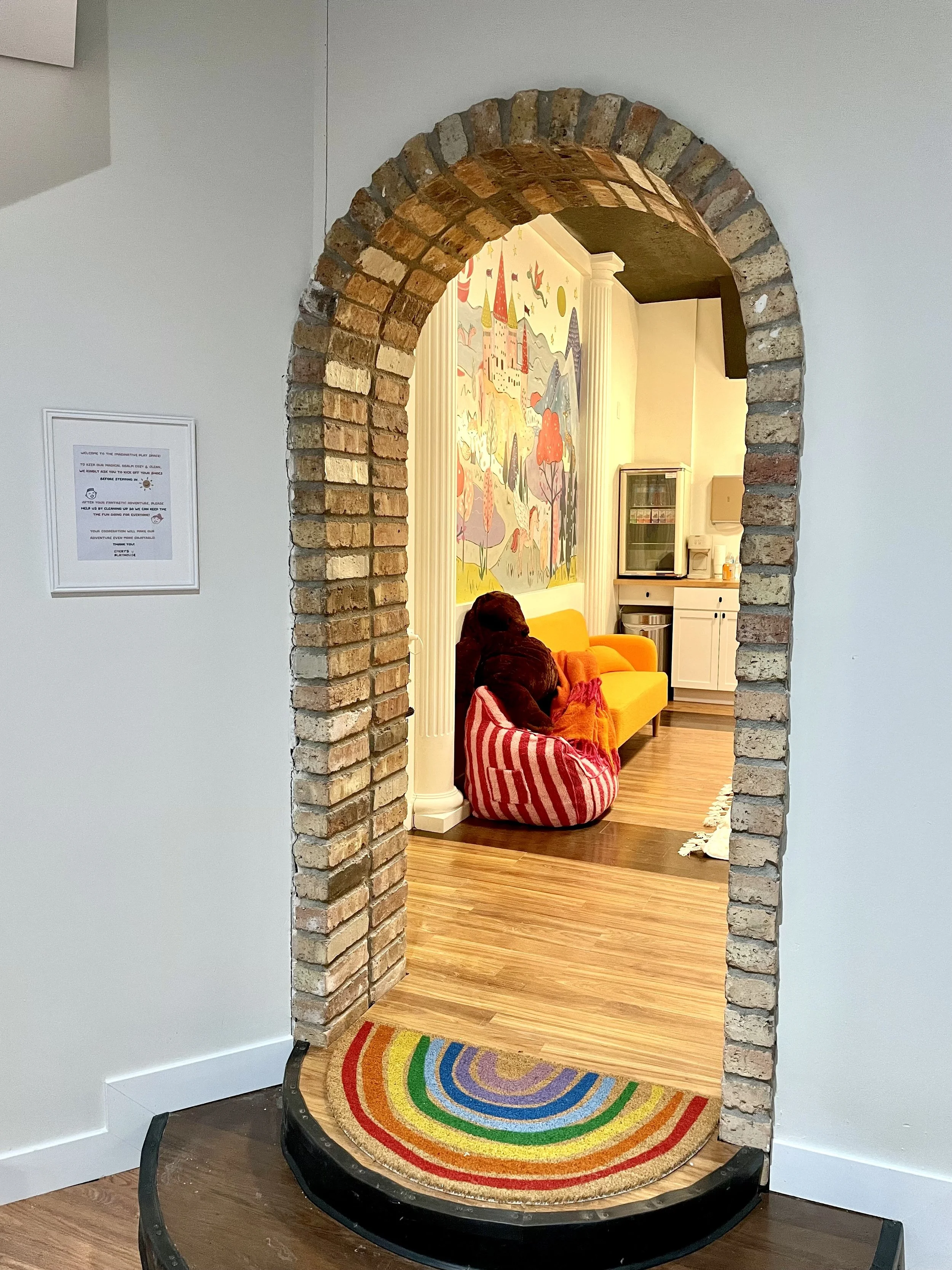 View through a brick archway into a colorful room with a yellow couch, a large stuffed bear, a rainbow mural, and wooden flooring.
