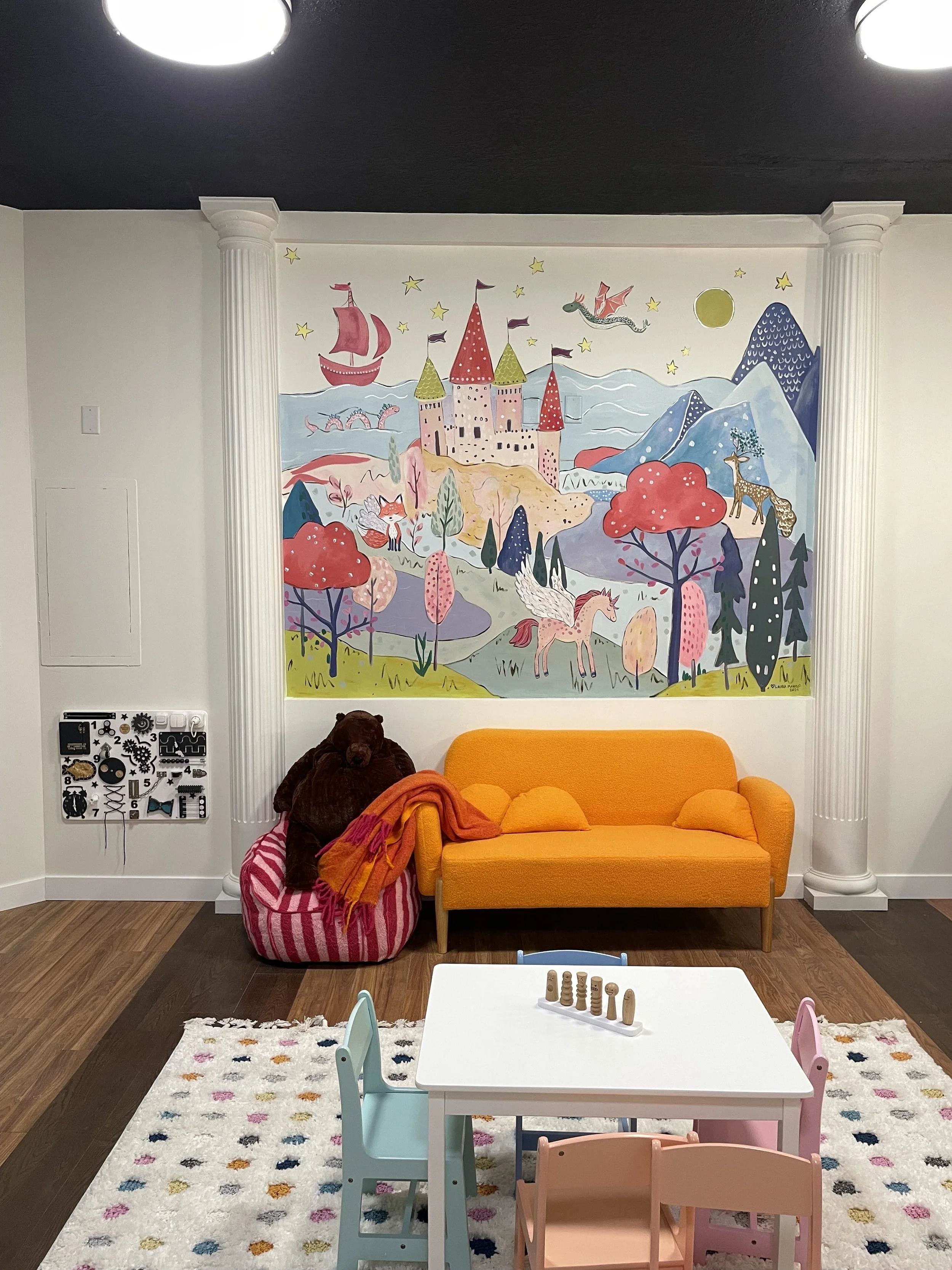 Colorful mural mural of a fairy tale castle with pink and green towers, trees, a fox, a unicorn with wings, a dragon, a giraffe, mountains, clouds, stars, and a red ship on the ocean. In front of the mural is a yellow sofa, a stuffed bear on a striped pink and white bean bag, a small white table with wooden game pieces, and three children-sized chairs in pastel pink, blue, and peach. The room has white walls with white decorative columns and a dark ceiling.