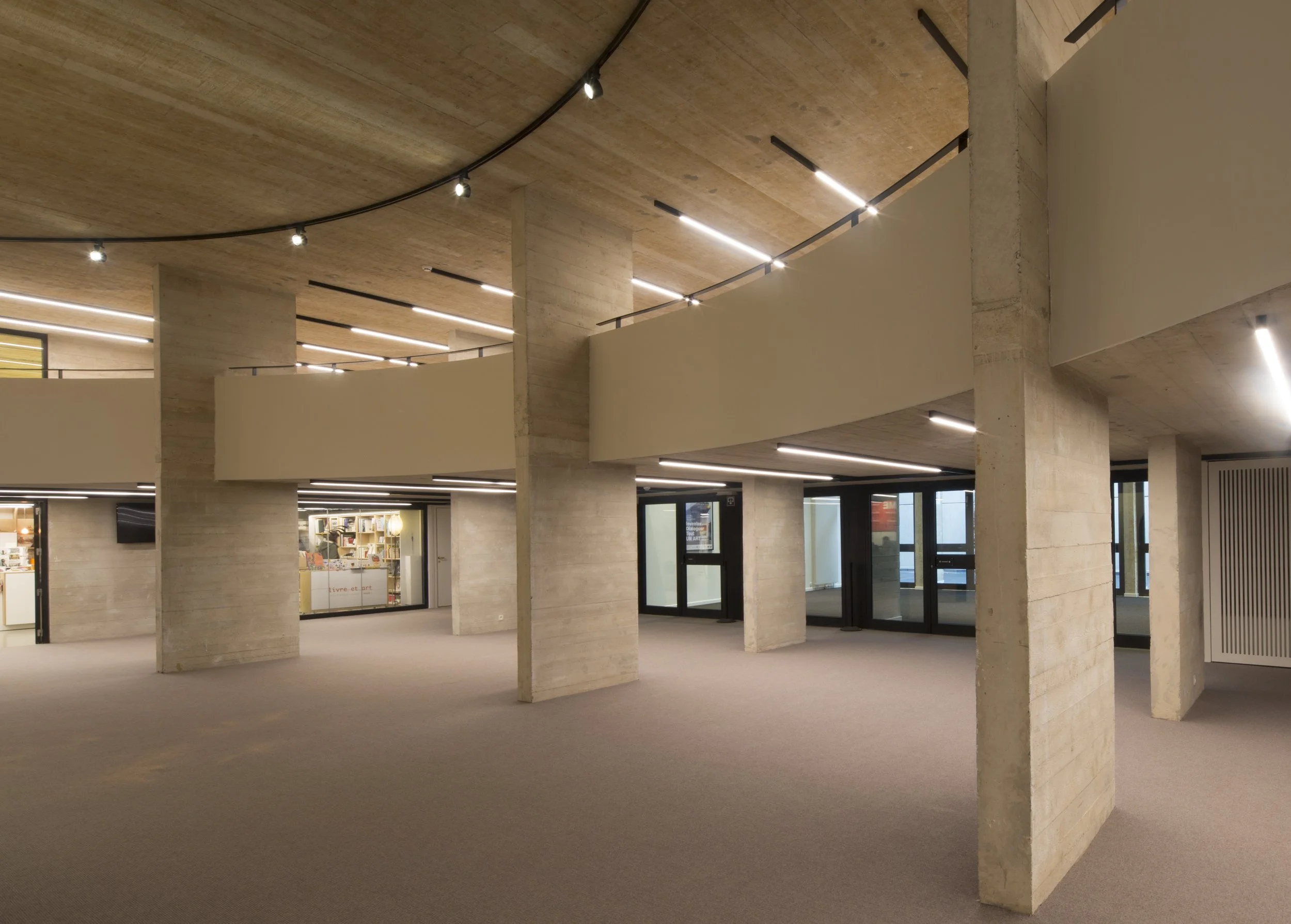 Axminster and Print Hospitality Carpets - suitable to pubs, clubs, hotels, bars:  Interior space with beige textured walls and ceiling, supported by concrete columns, illuminated by modern linear and spot lighting fixtures.