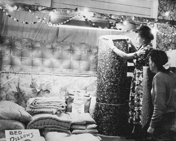 6o years of expertise: A woman and a girl are examining a large rolled-up quilt or blanket at a store display. The display includes stacks of pillows, quilts, and bed linens, with a sign that reads 'BED PILLOWS'.