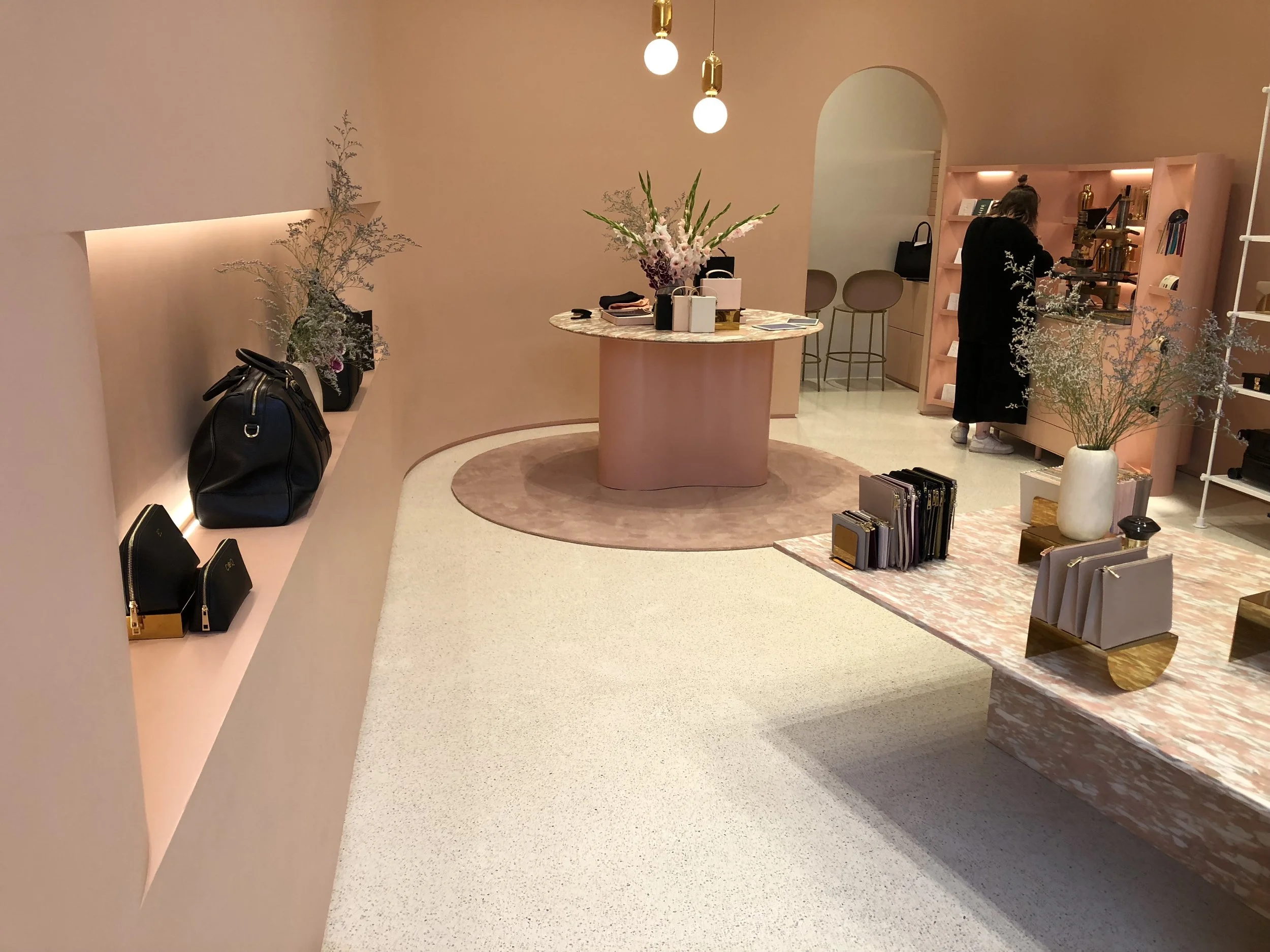 Inside a boutique or showroom with pastel pink walls and modern decor. A marble table in the center holds flowers and shopping bags. Shopping bags and handbags are displayed on the curved wall to the left. A person is browsing items on a pink shelvin