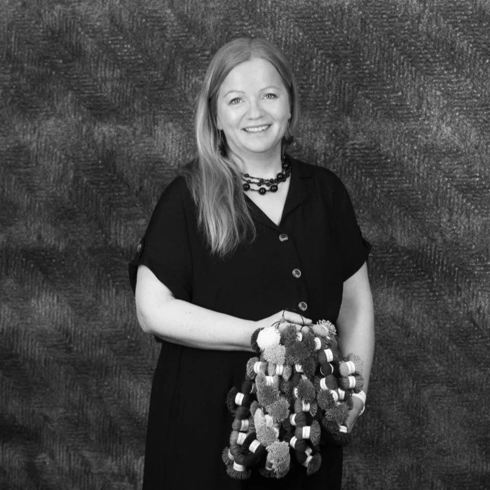 A woman with long hair wearing a black dress and a beaded necklace, holding a bunch of colorful yarn balls, standing in front of a textured wall.