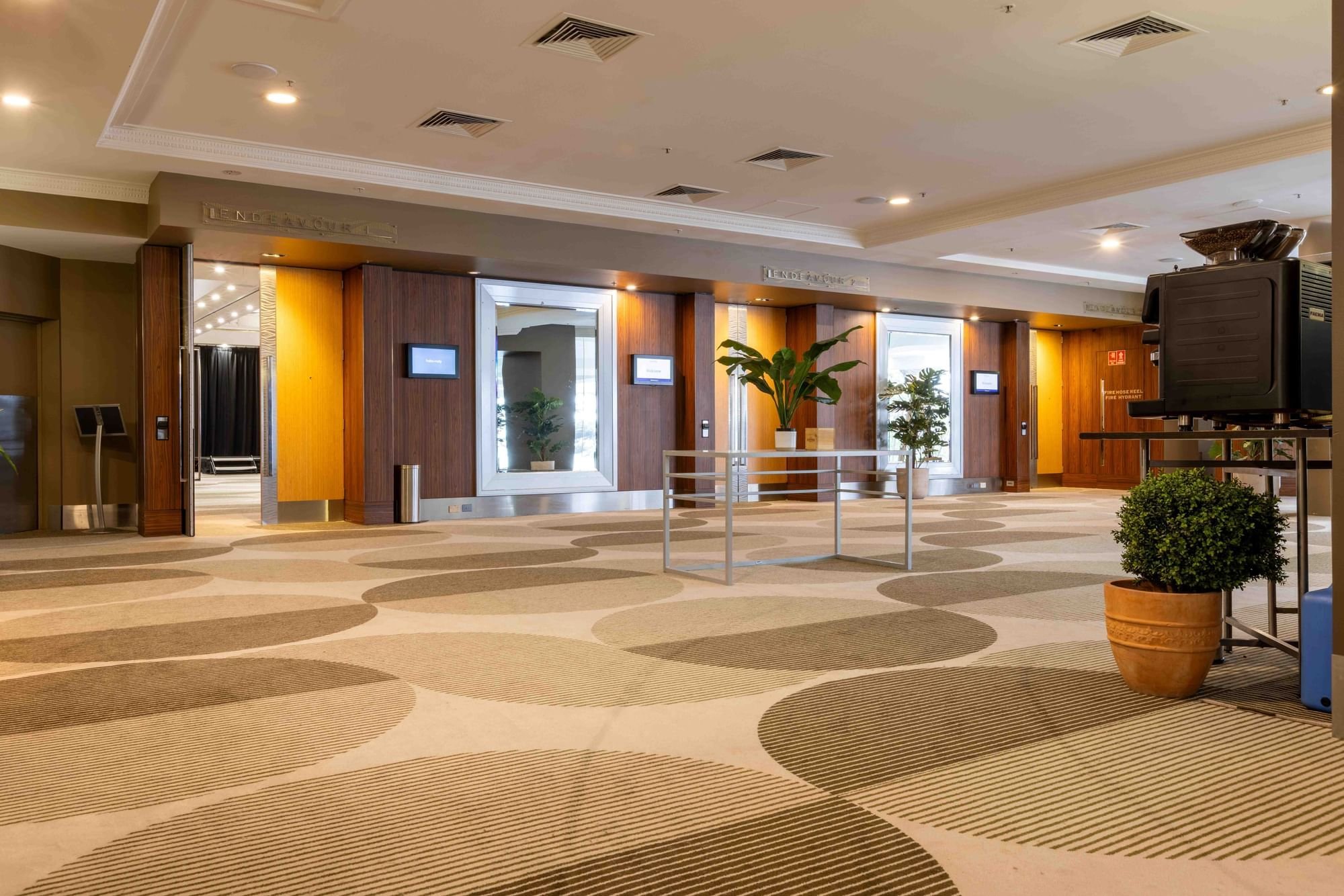 Axminster and Print Hospitality Carpets - suitable to pubs, clubs, hotels, bars, gaming: Interior of a hotel lobby with carpeted floor, potted plants, elevators, and wall-mounted screens, illuminated by ceiling lights.