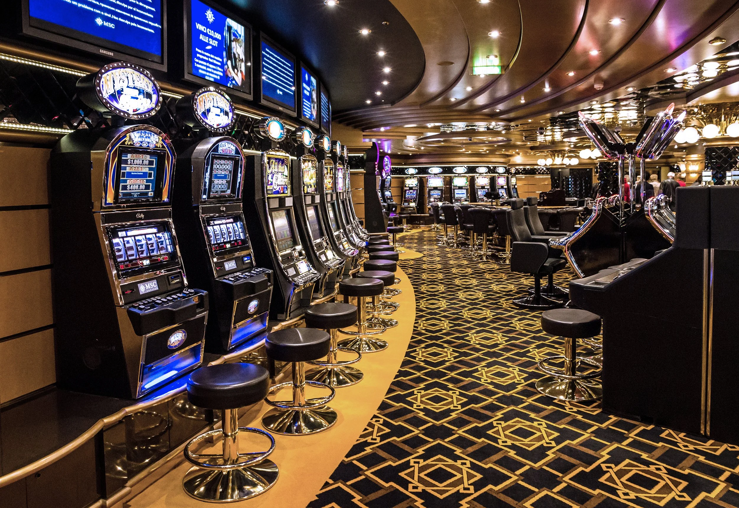 Axminster and Print Hospitality Carpets - suitable to pubs, clubs, hotels, bars:  Interior of a casino with row of slot machines and bar stools, patterned carpet, digital screens on the wall, and a group of people in the background.