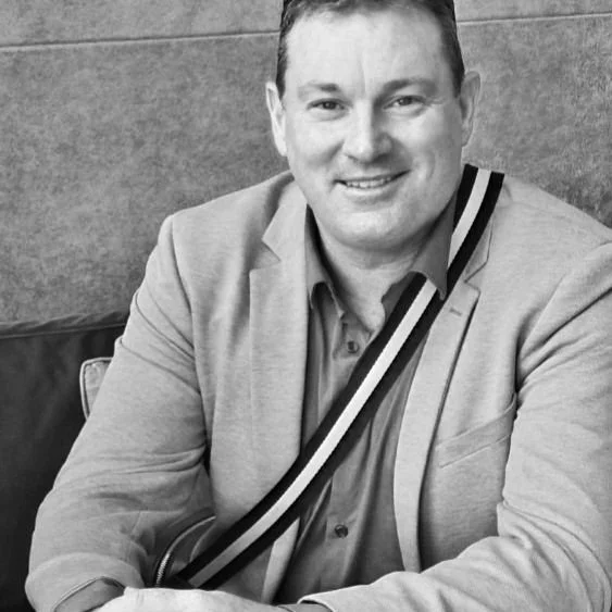 A smiling man in a suit jacket sitting at a table, with a striped bag strap over his shoulder, in an indoor setting.