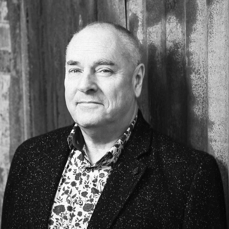Black and white portrait of a middle-aged man with short hair, wearing a patterned shirt and a speckled blazer, standing against a wooden wall.