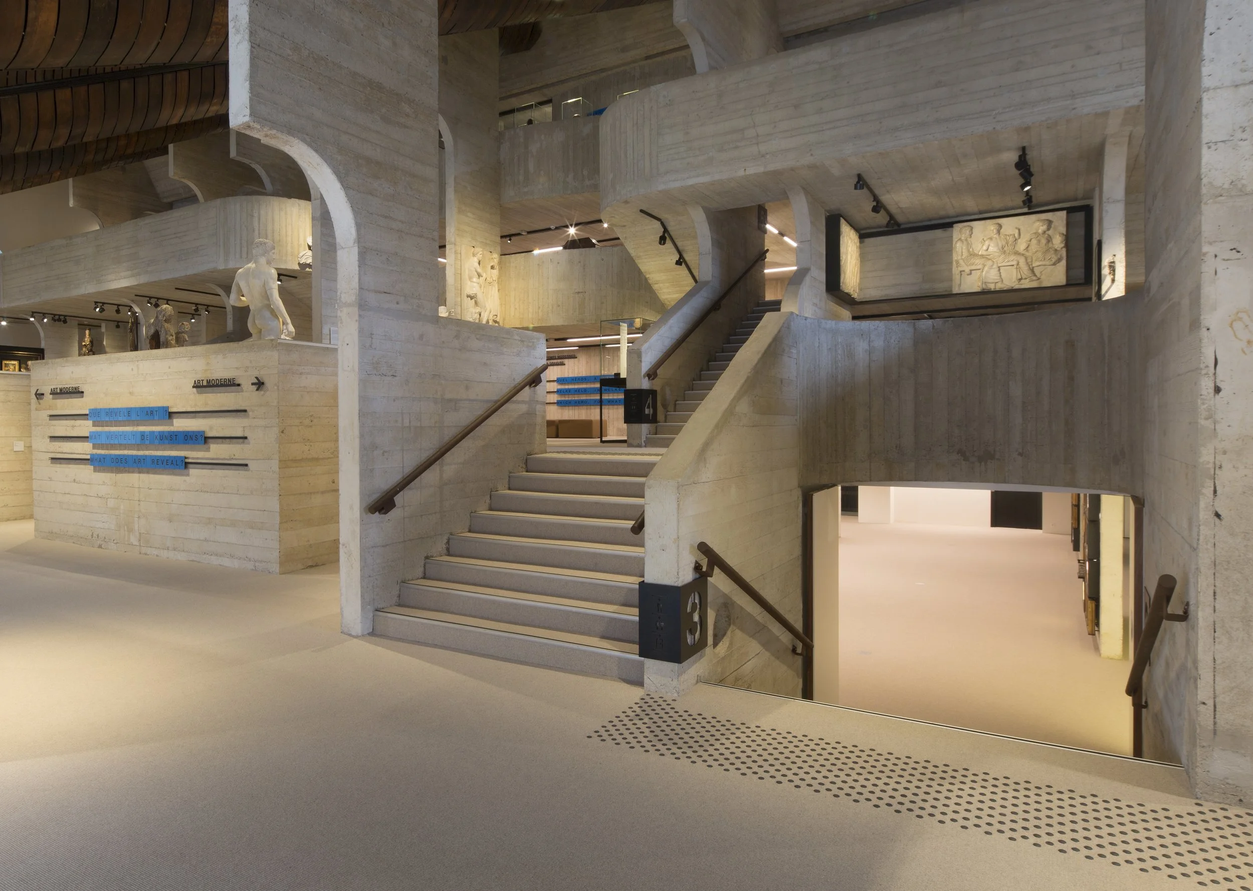 Axminster and Print Hospitality Carpets - suitable to pubs, clubs, hotels, bars:  Interior of an art museum with concrete walls and stairs, displaying sculptures and wall art, and directional signs.