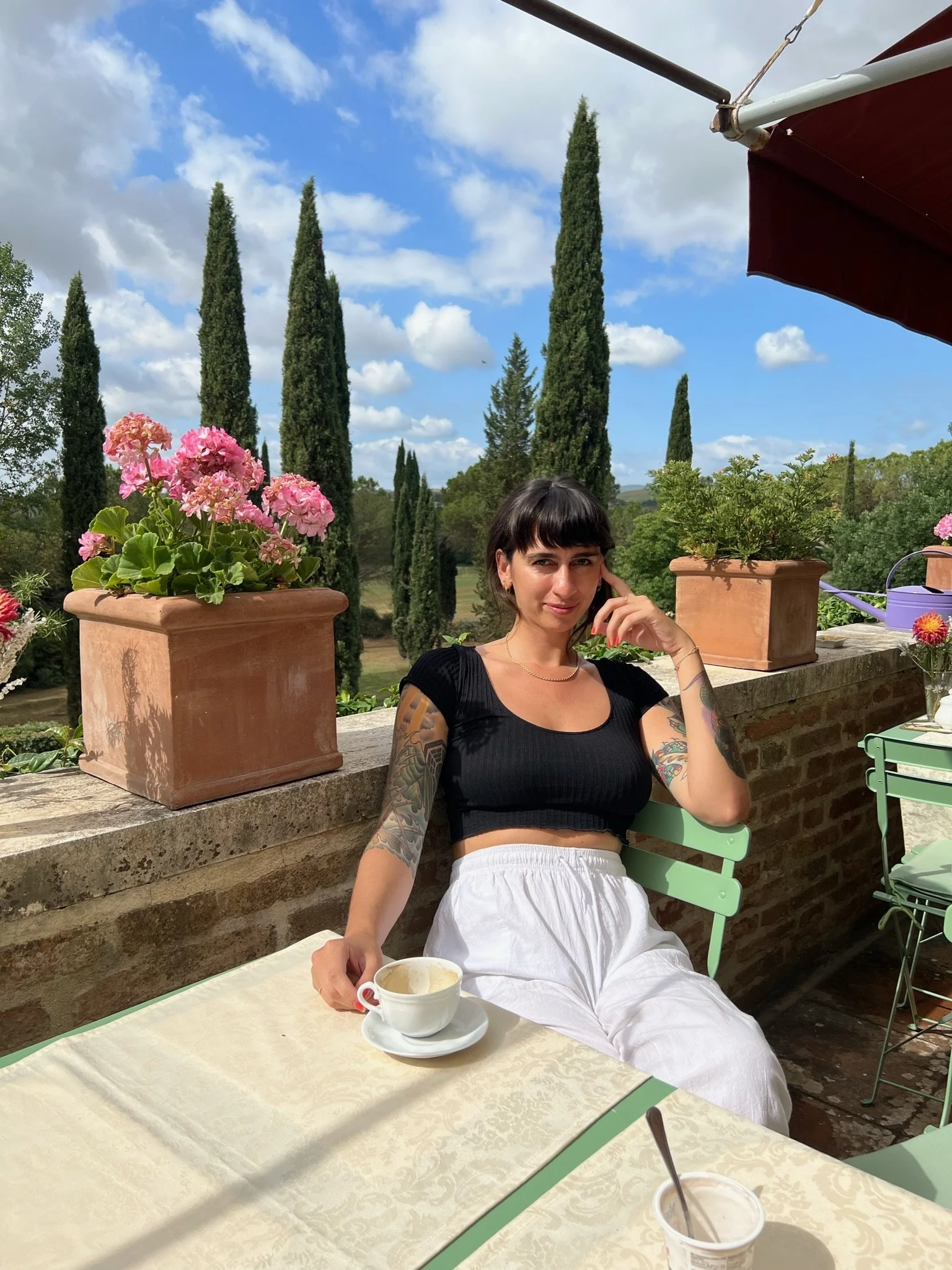 A woman with short dark hair and tattoos on her arms, sitting at an outdoor table on a terrace with potted pink and red flowers behind her. She is wearing a black crop top and white pants, holding a cup of coffee or tea, with a sunny sky and tall cypress trees in the background.