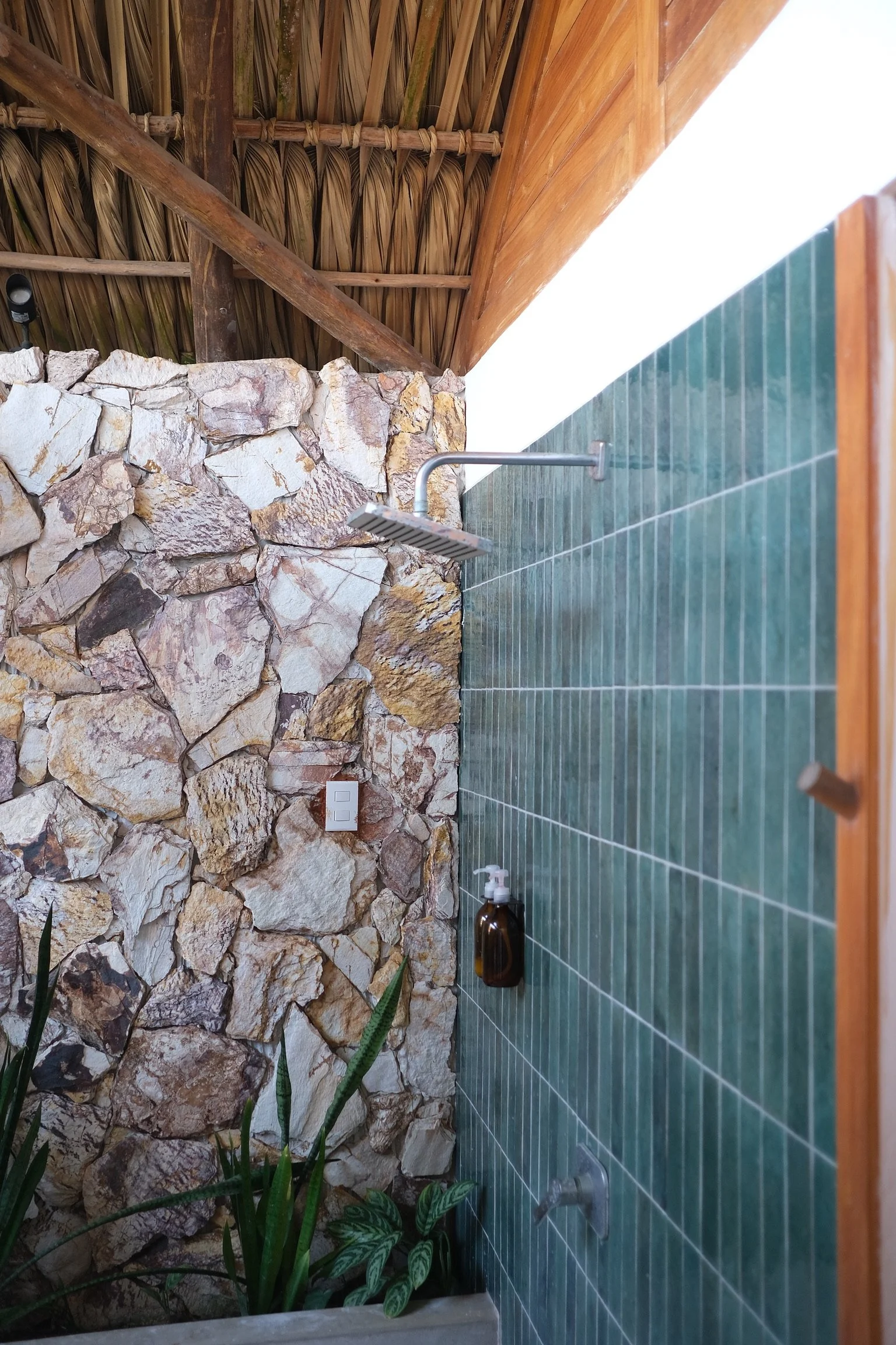 Outdoor shower with a green-tiled wall, a rustic stone wall, a wooden roof, and plants at the base.