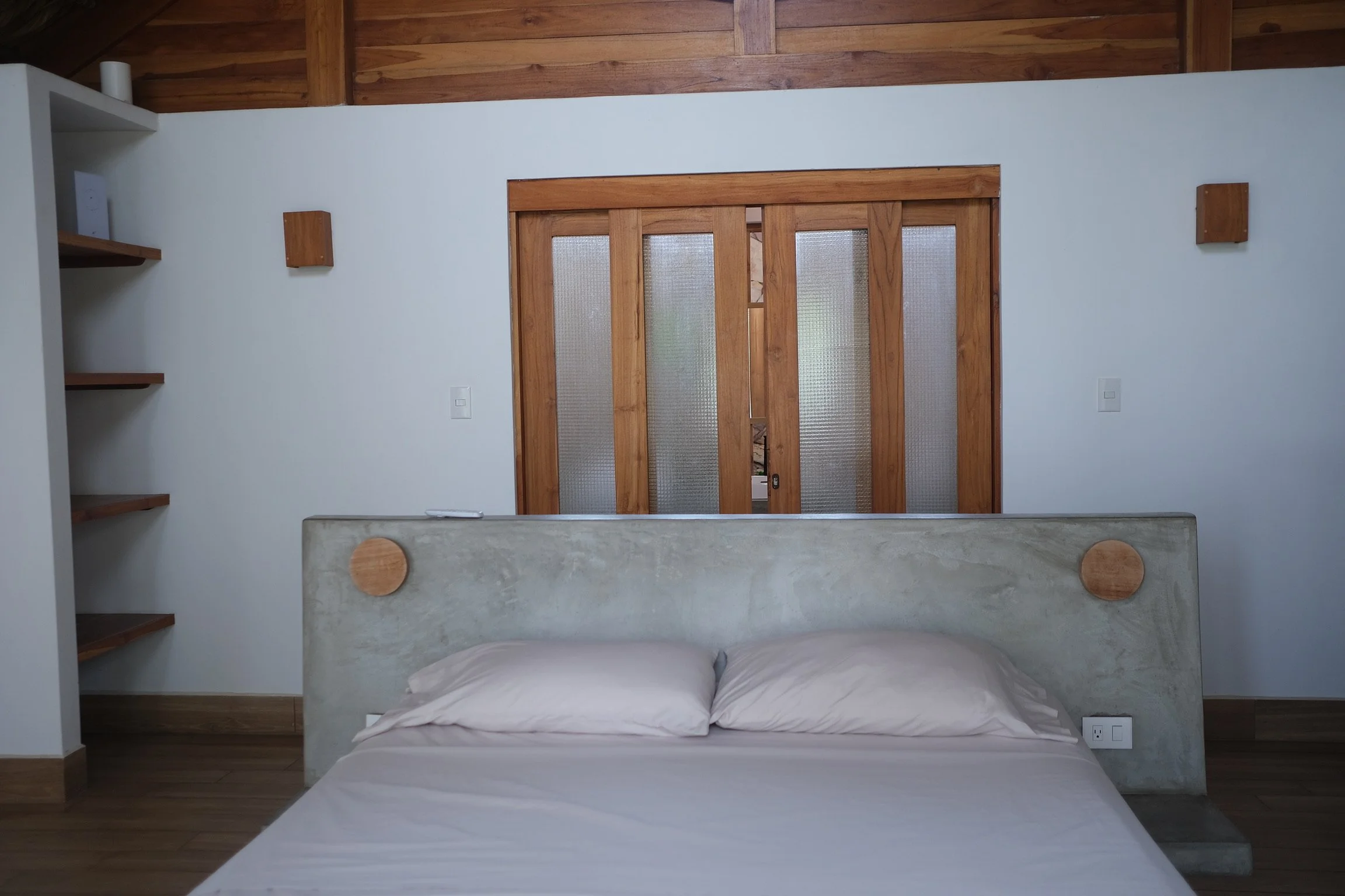 A minimalist bedroom with a concrete bed frame, beige pillows, and a wooden sliding door with frosted glass panels, against a white wall with wooden accents.