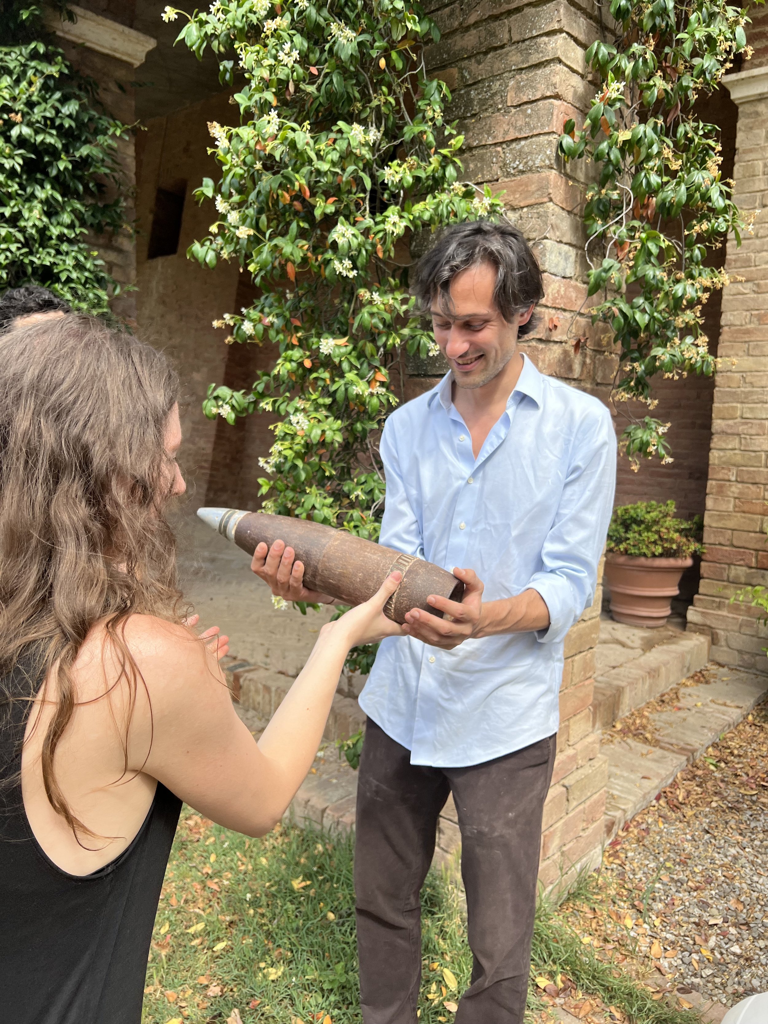 A man in a light blue shirt and a woman with long brown hair exchanging a large, cylindrical object resembling a rocket or missile outside near a brick building with greenery.