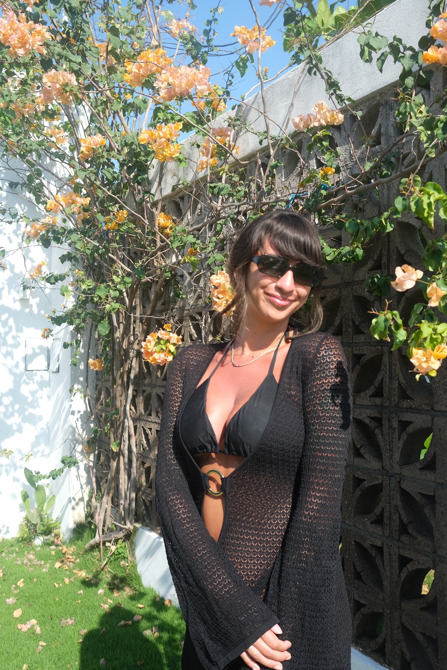 A woman wearing sunglasses and a black bikini top with a black open-knit cover-up standing outdoors near a flowering vine and a concrete block wall.