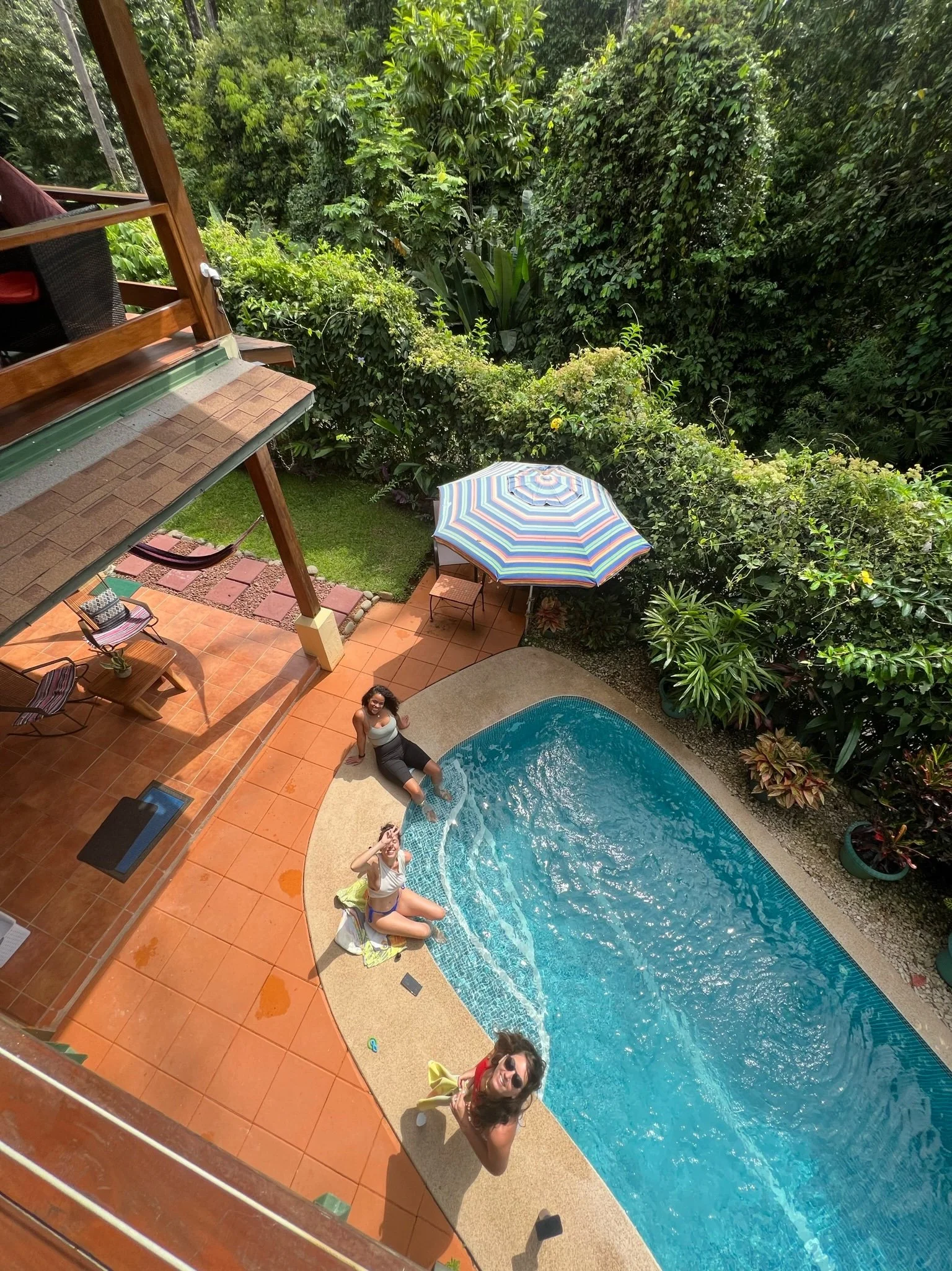 A backyard with a swimming pool, three women sitting and relaxing by the pool, surrounded by lush green trees and plants, a patio with outdoor furniture, an umbrella, and a grassy area.