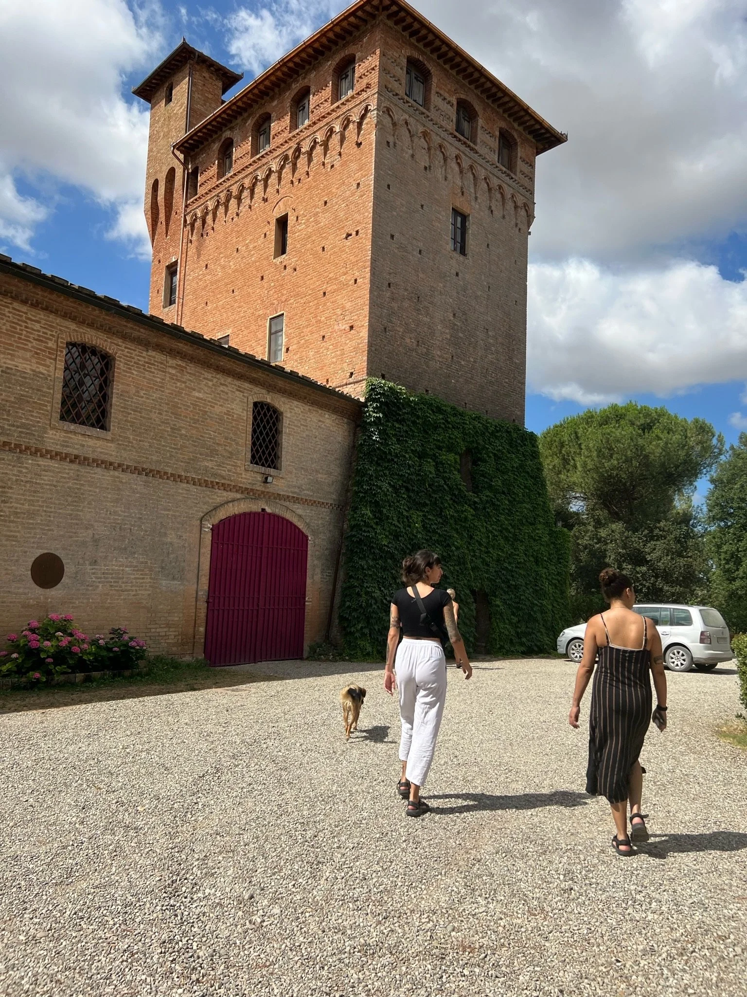 Two women walking toward a medieval brick castle with a tower, surrounded by trees and a gravel path, under partly cloudy skies.