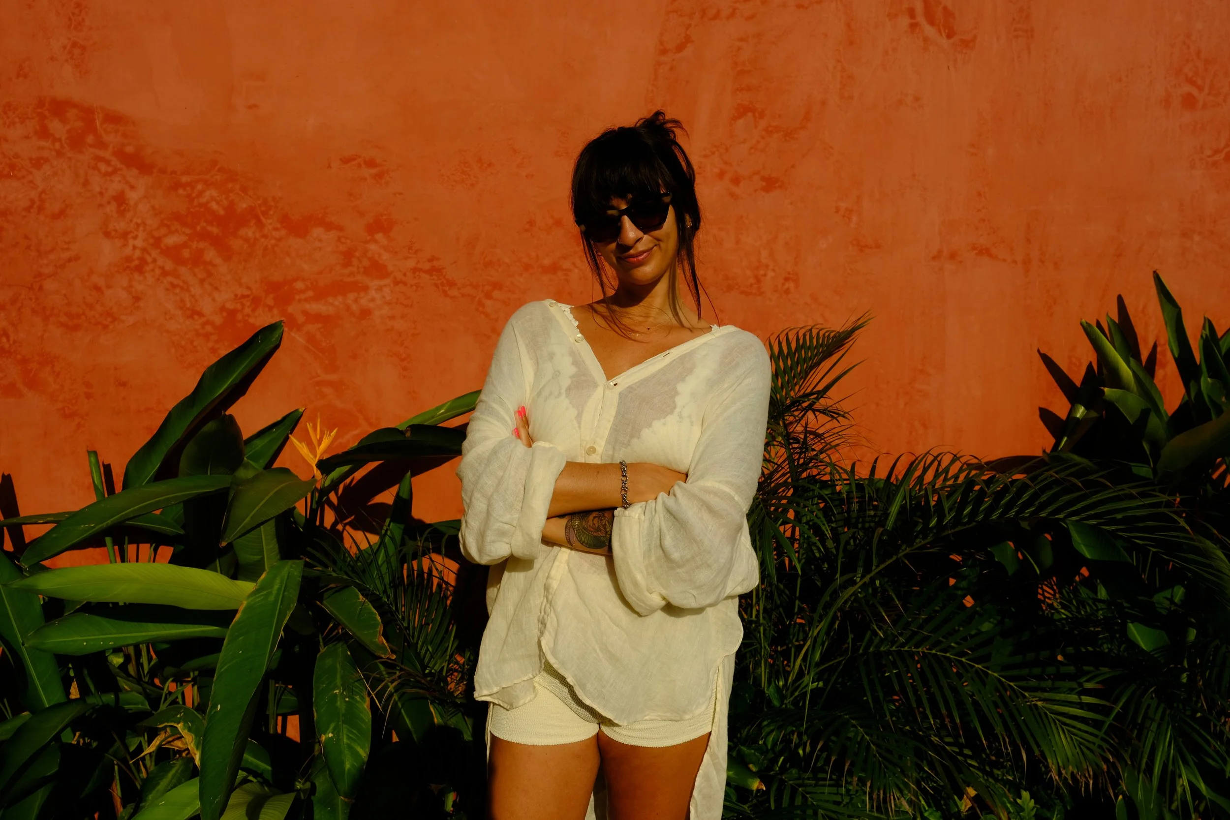 A woman wearing sunglasses and a light-colored, loose shirt with rolled-up sleeves, standing with arms crossed among green tropical plants against an orange textured wall.