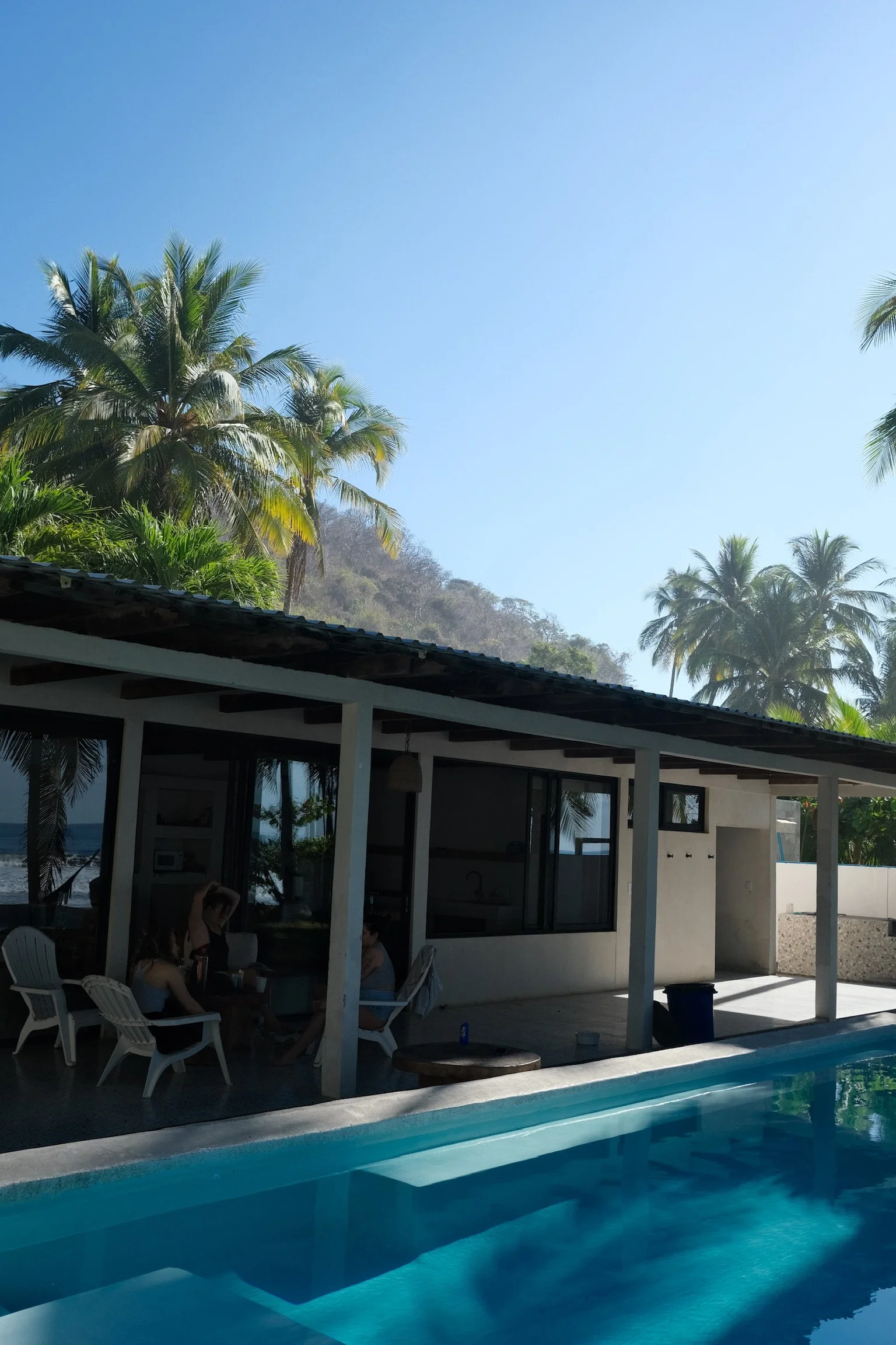 People relaxing by a pool under a shaded patio, with palm trees and a clear blue sky in the background at a tropical location.