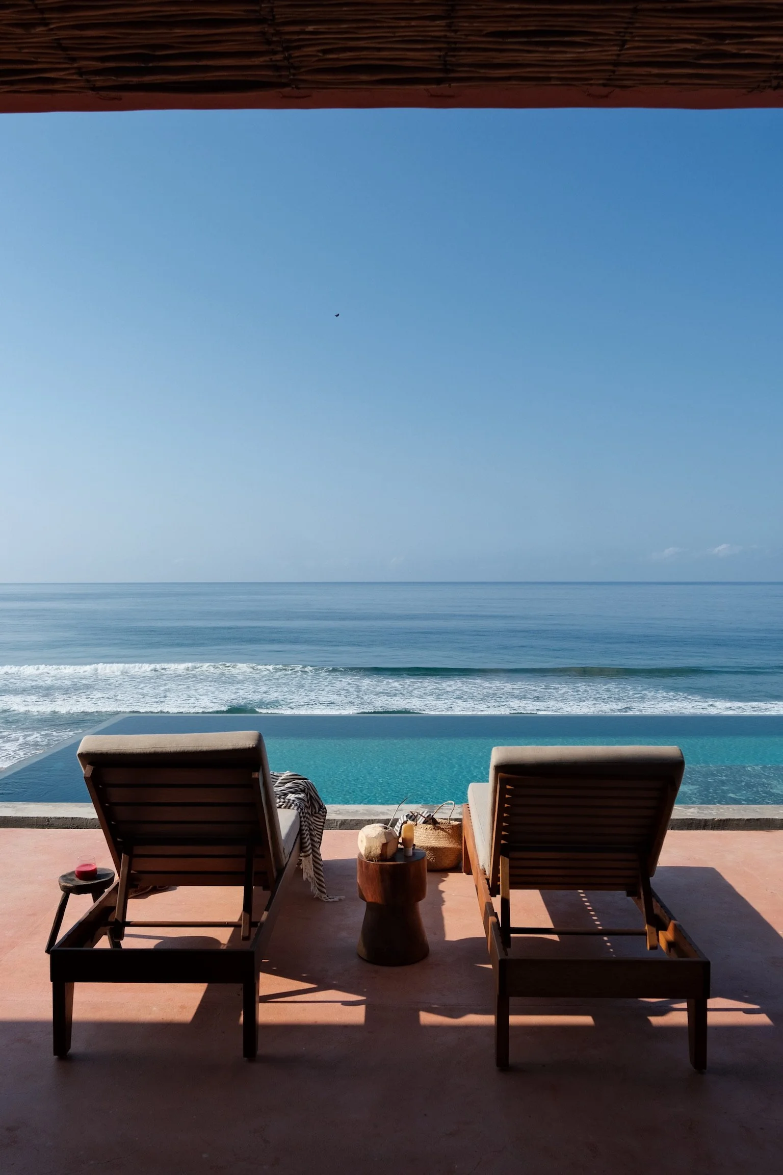 Two lounge chairs on a patio facing an infinity pool and ocean, with a clear blue sky overhead.